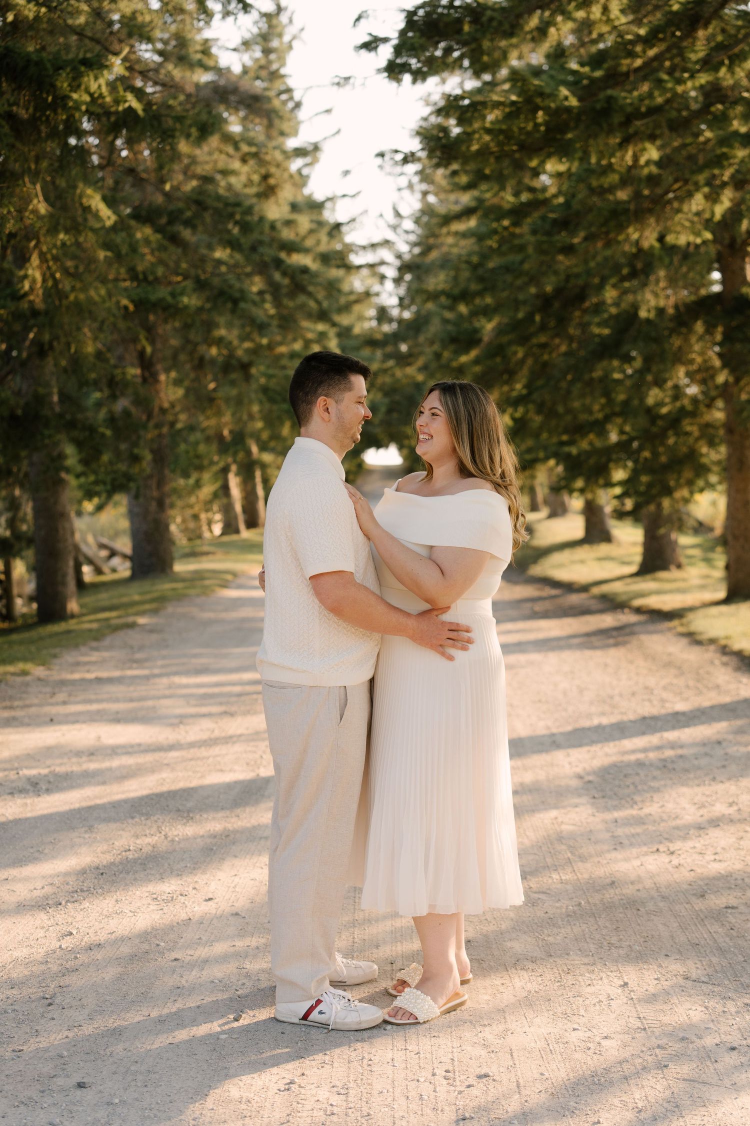 Smiling couple in matching cream outfits embracing on a sunlit tree-lined gravel path in summer.