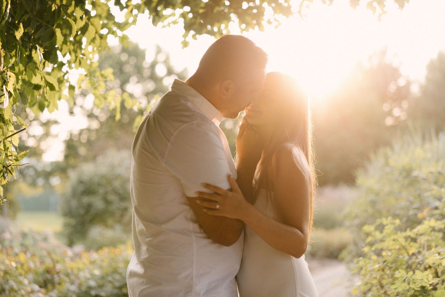 Couple kissing in a lush garden with warm golden sunset light glowing through surrounding green trees.
