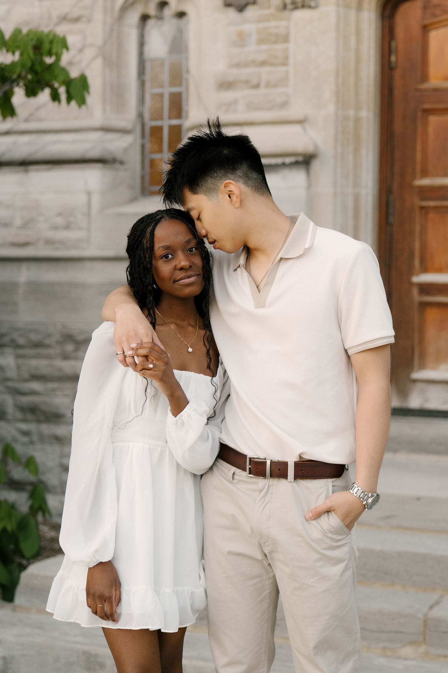 Interracial couple in white outfits posing together on steps of a stone building.