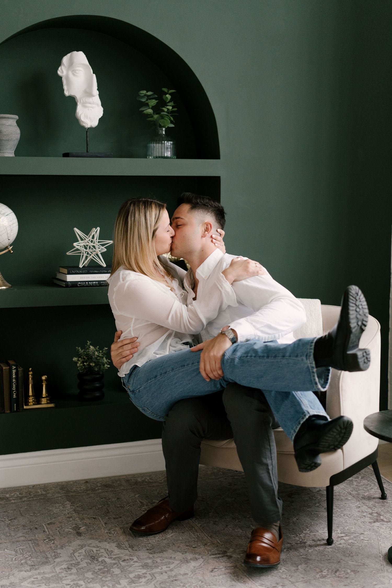 Couple sharing a passionate kiss while sitting together in a cream chair in a green-walled room.