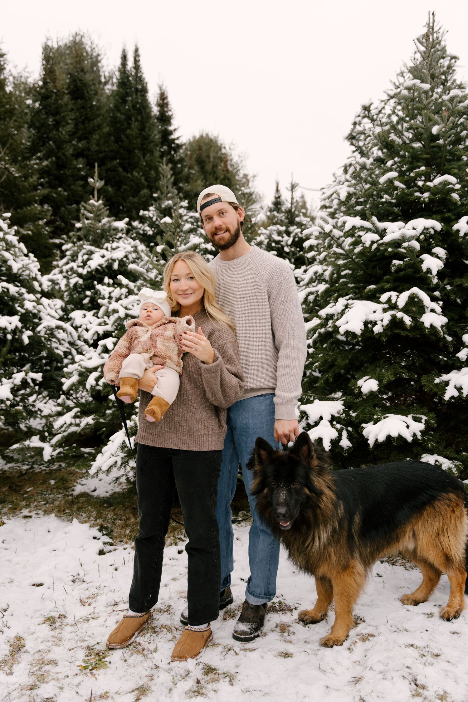 Young family with baby and German Shepherd dog posing among snow-covered pine trees in winter.