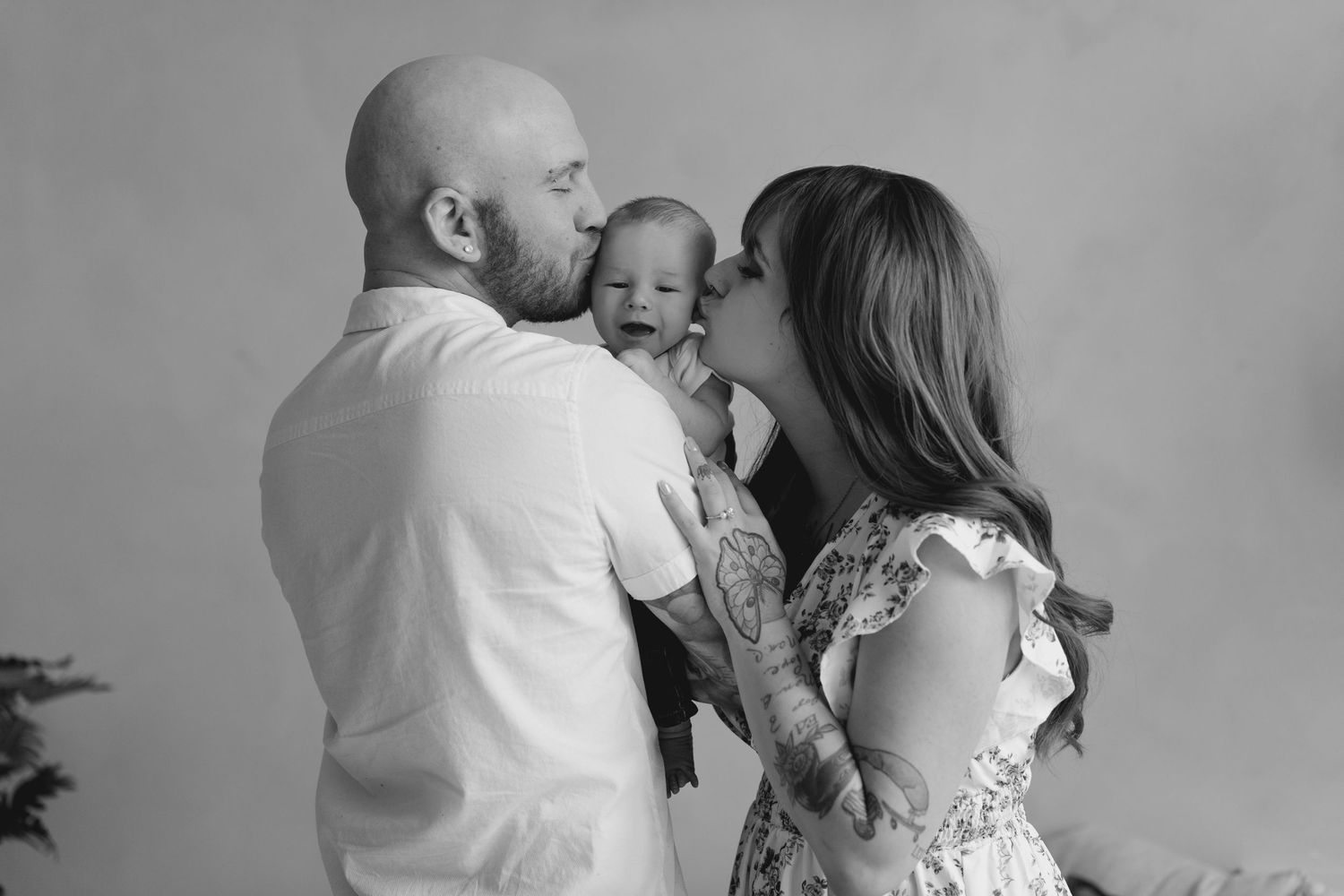 Black and white photo of tattooed parents kissing their smiling newborn baby on each cheek.