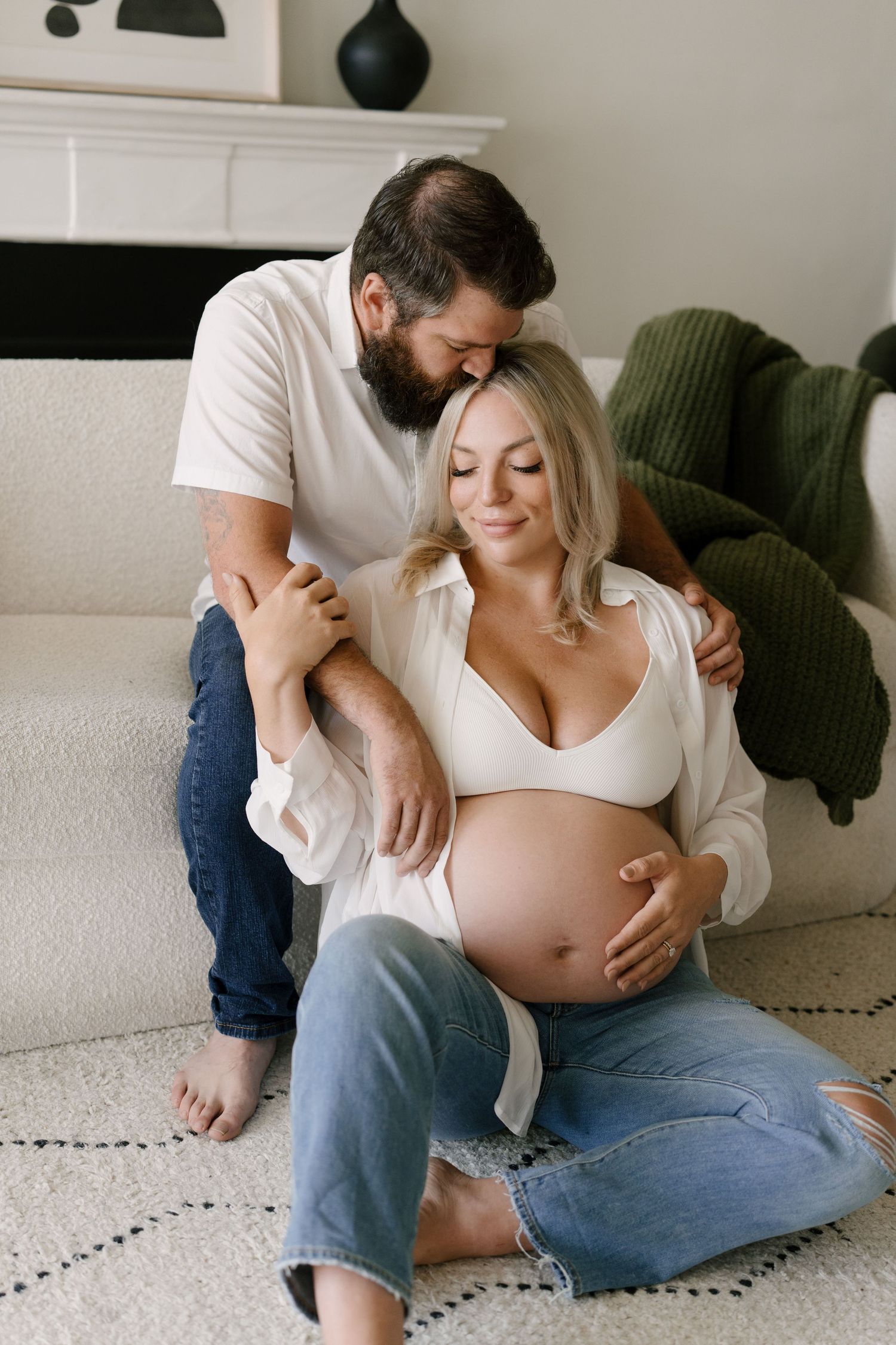 A bearded man tenderly embraces his pregnant partner as they sit together on a cozy living room floor.