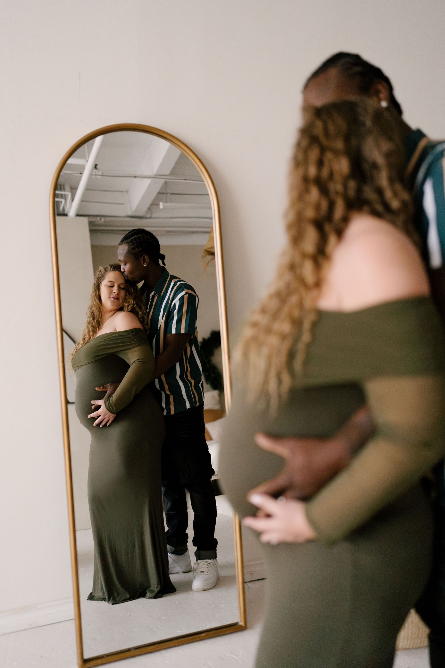 Interracial couple reflected in gold arch mirror, pregnant woman in olive dress during maternity shoot.