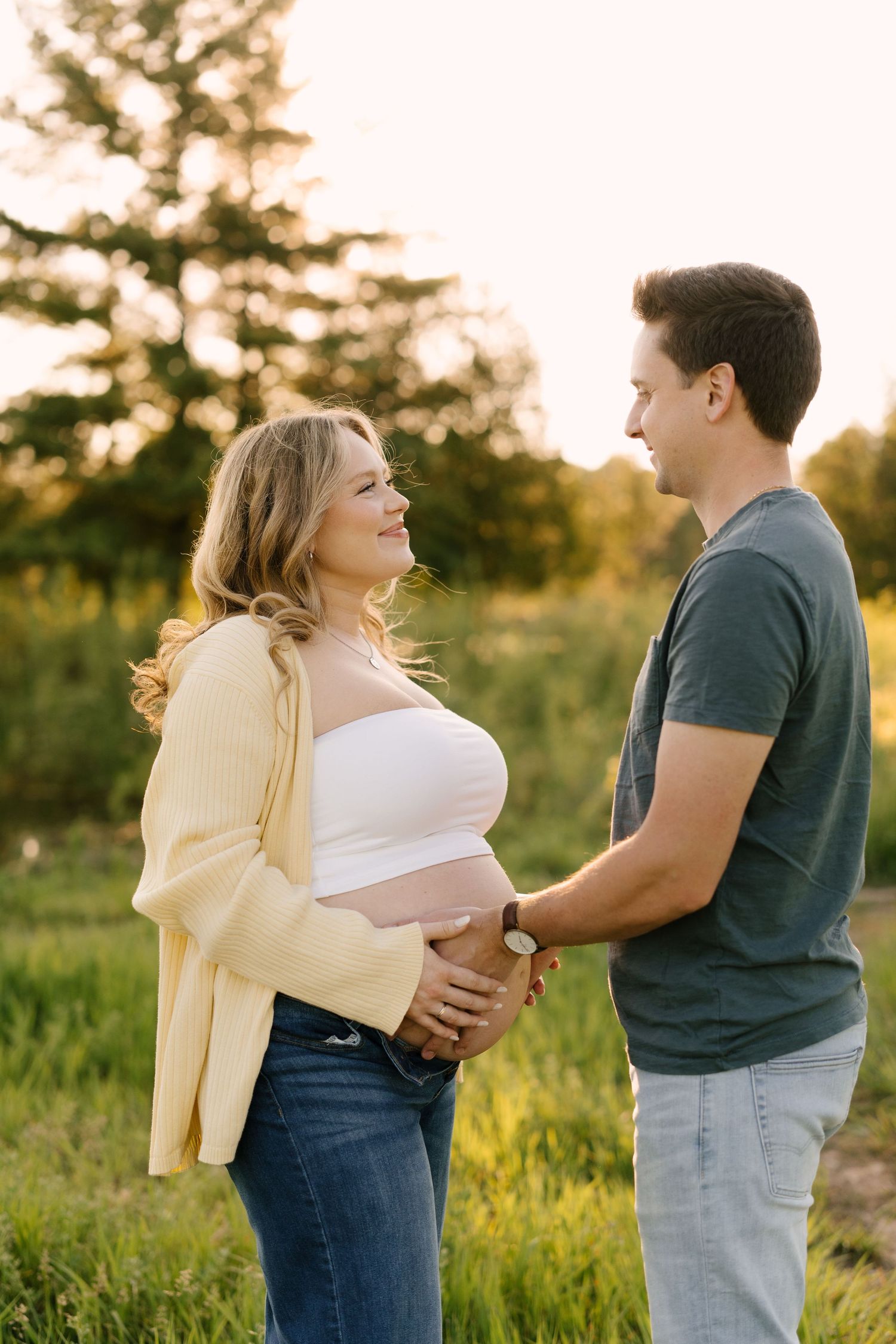 Couple facing each other outdoors at golden hour, both holding pregnant womans baby bump in a field.