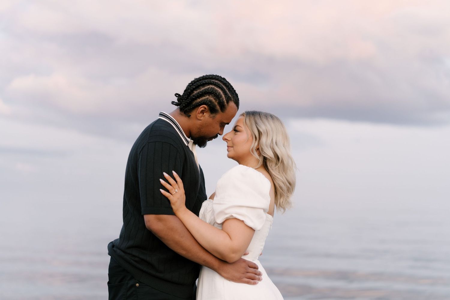 An interracial couple leans in for a kiss while embracing on a beach with a pastel cloudy sky behind them.