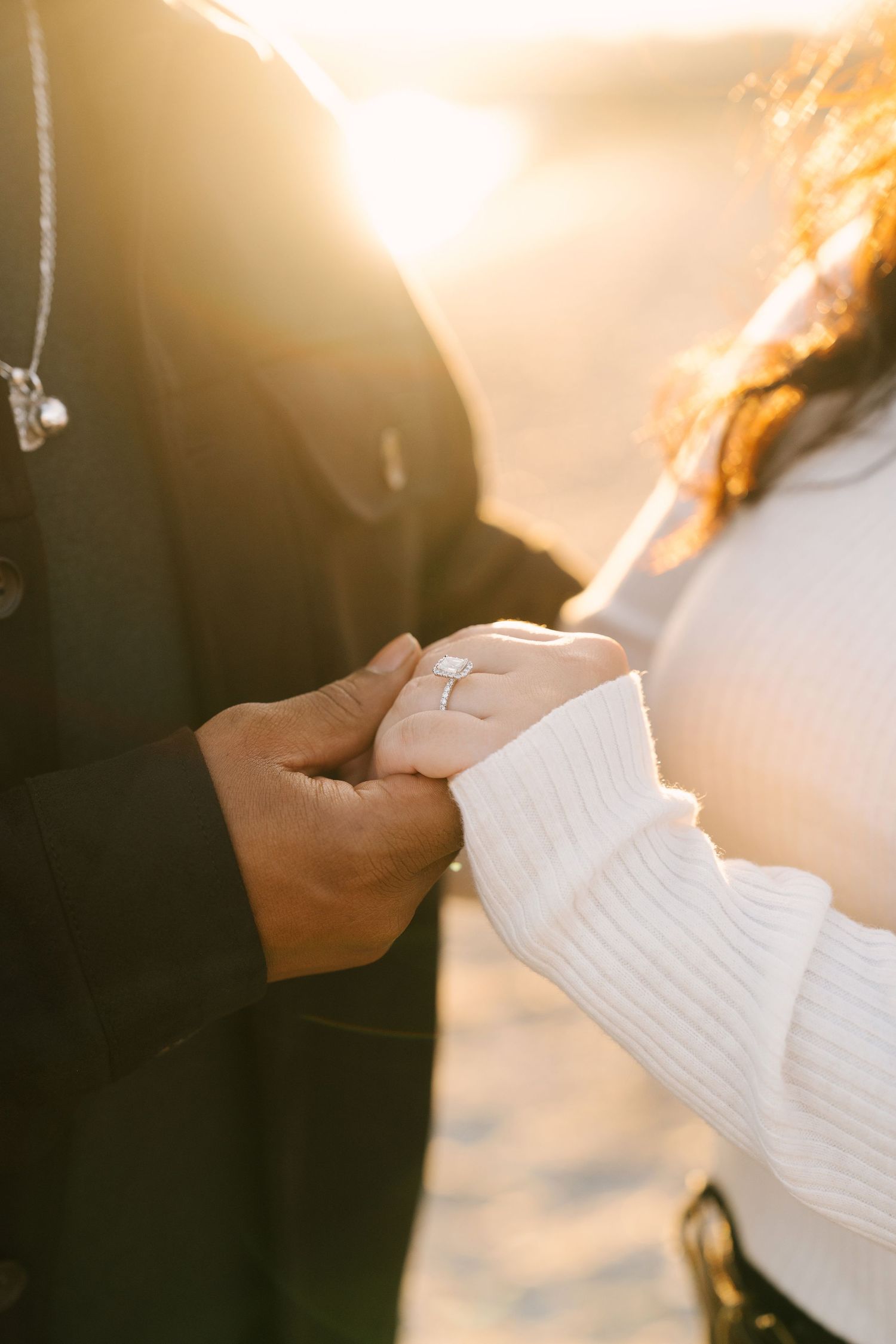Close-up of couple holding hands at sunset, woman wearing an engagement ring on the beach.