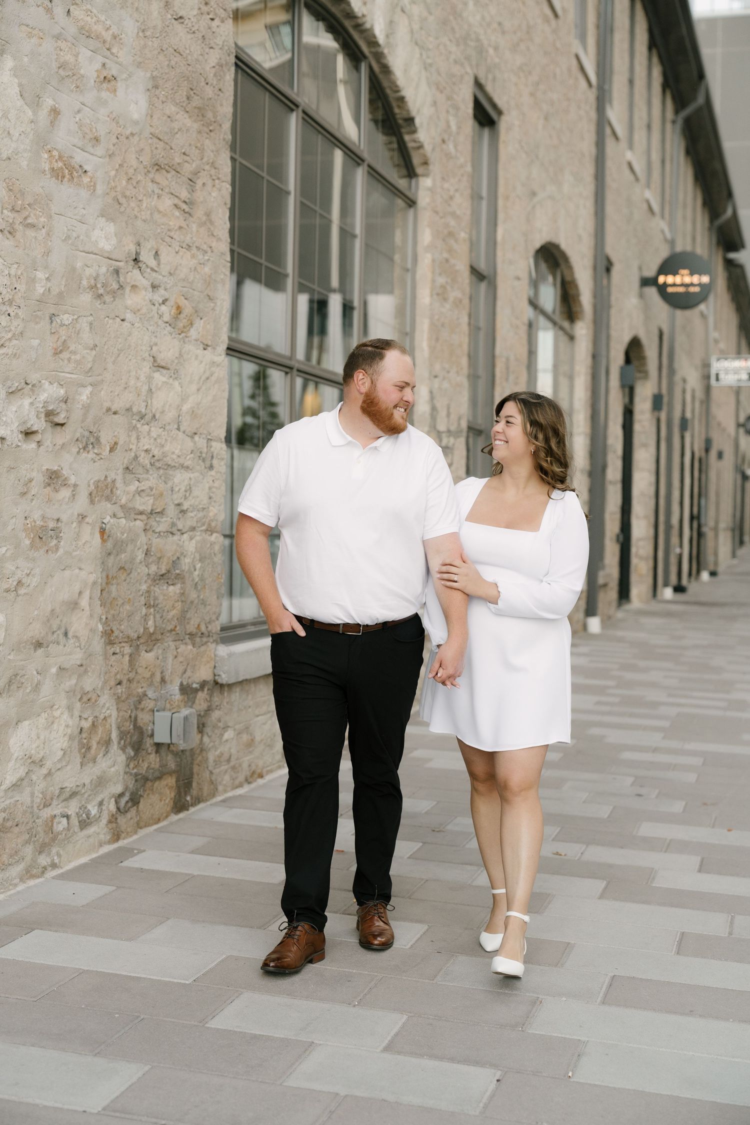 Couple in matching white outfits walking and smiling at each other along a stone building pathway.