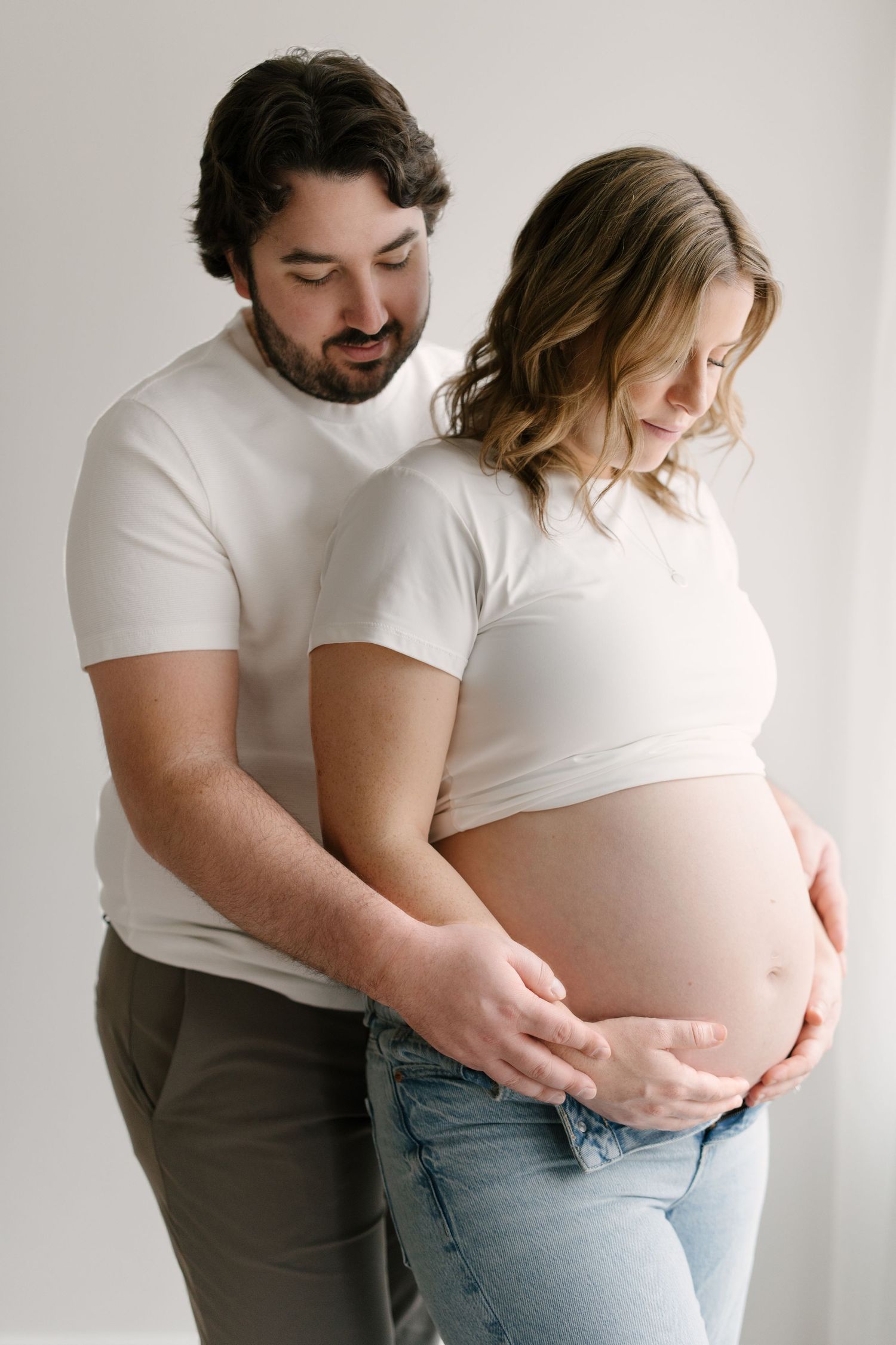 A couple in white tops lovingly cradling the pregnant woman's bare baby bump together.