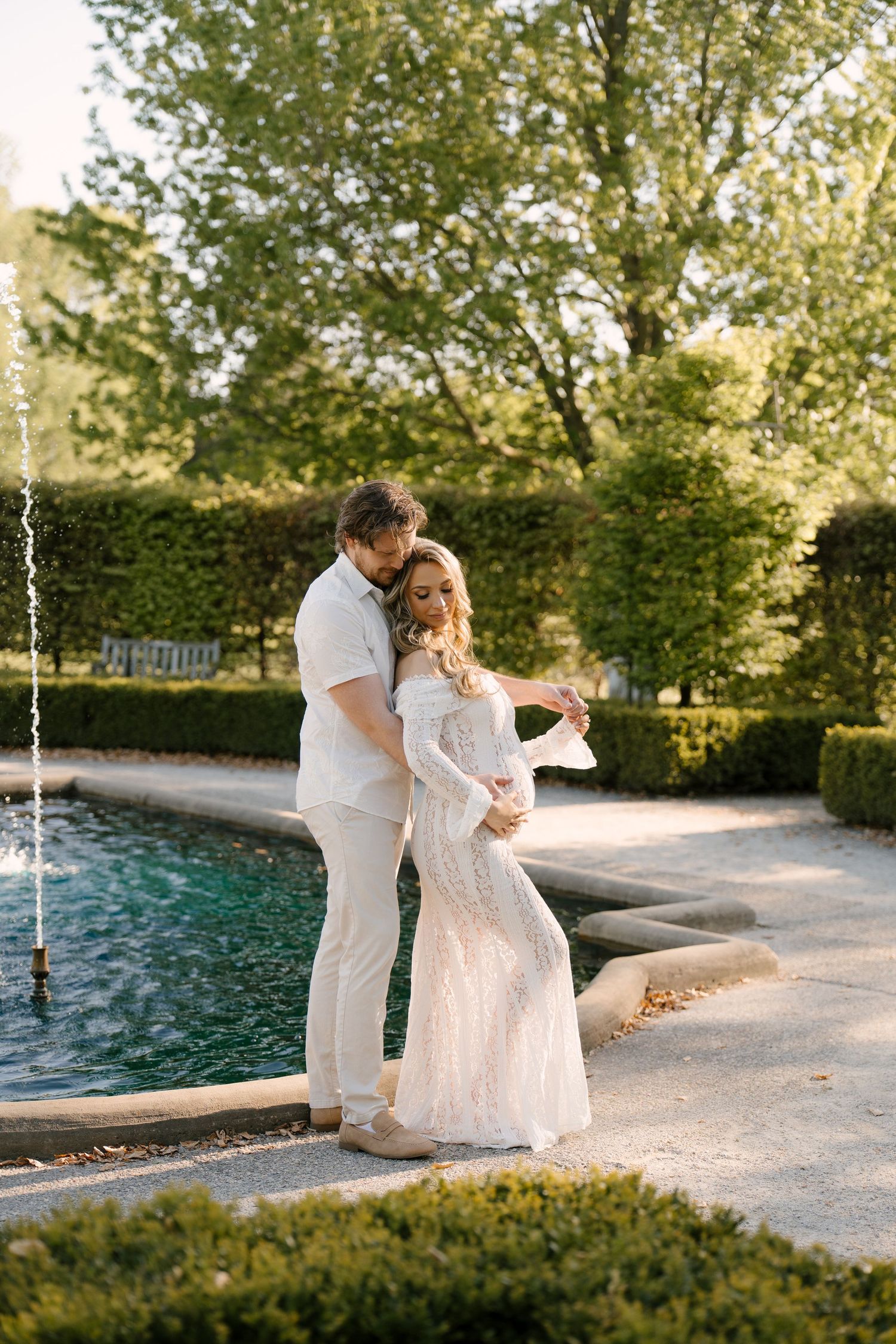 Couple in white outfits embracing by a garden fountain surrounded by lush green trees during golden hour.
