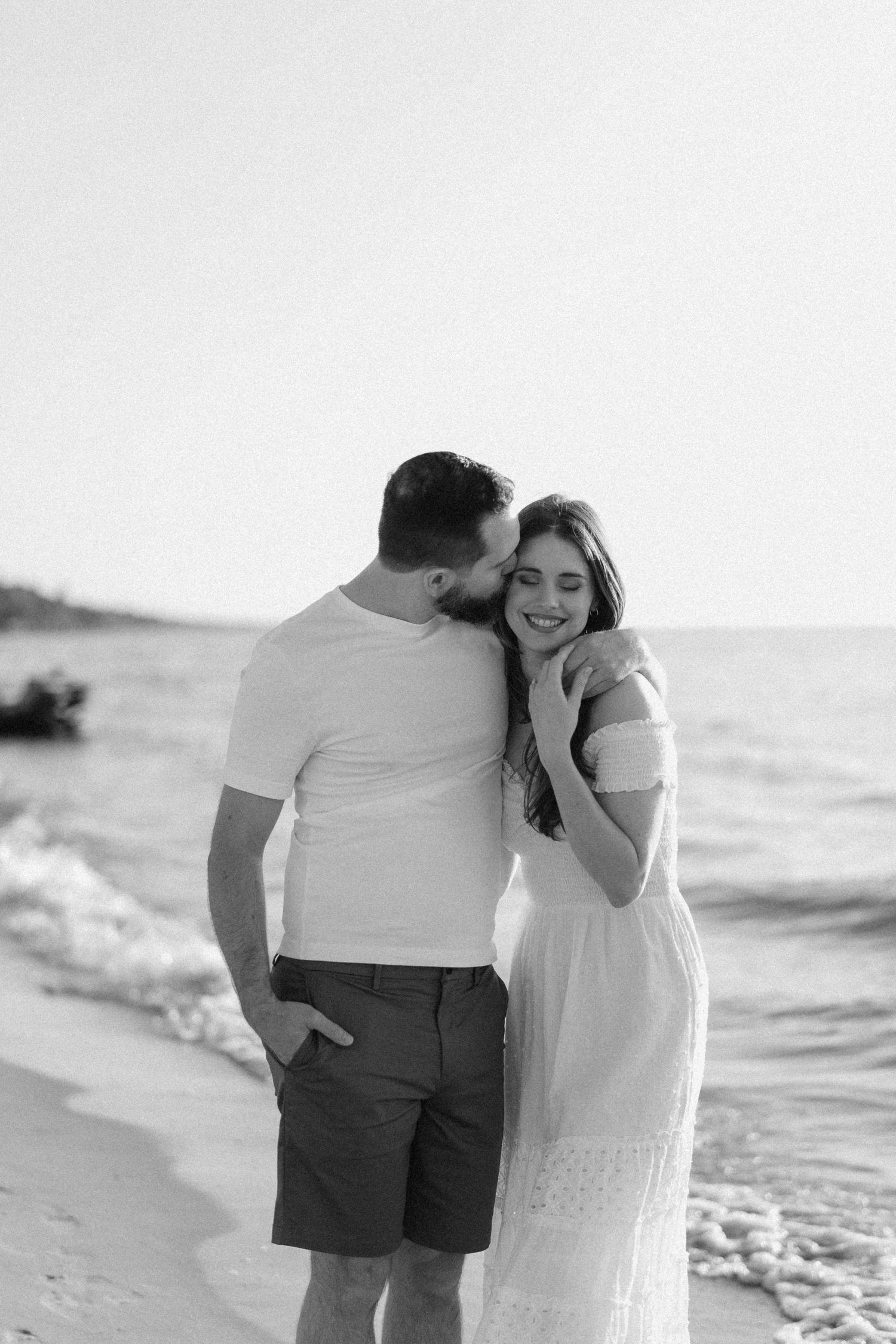 Black and white photo of smiling couple embracing on a sandy beach by the ocean.