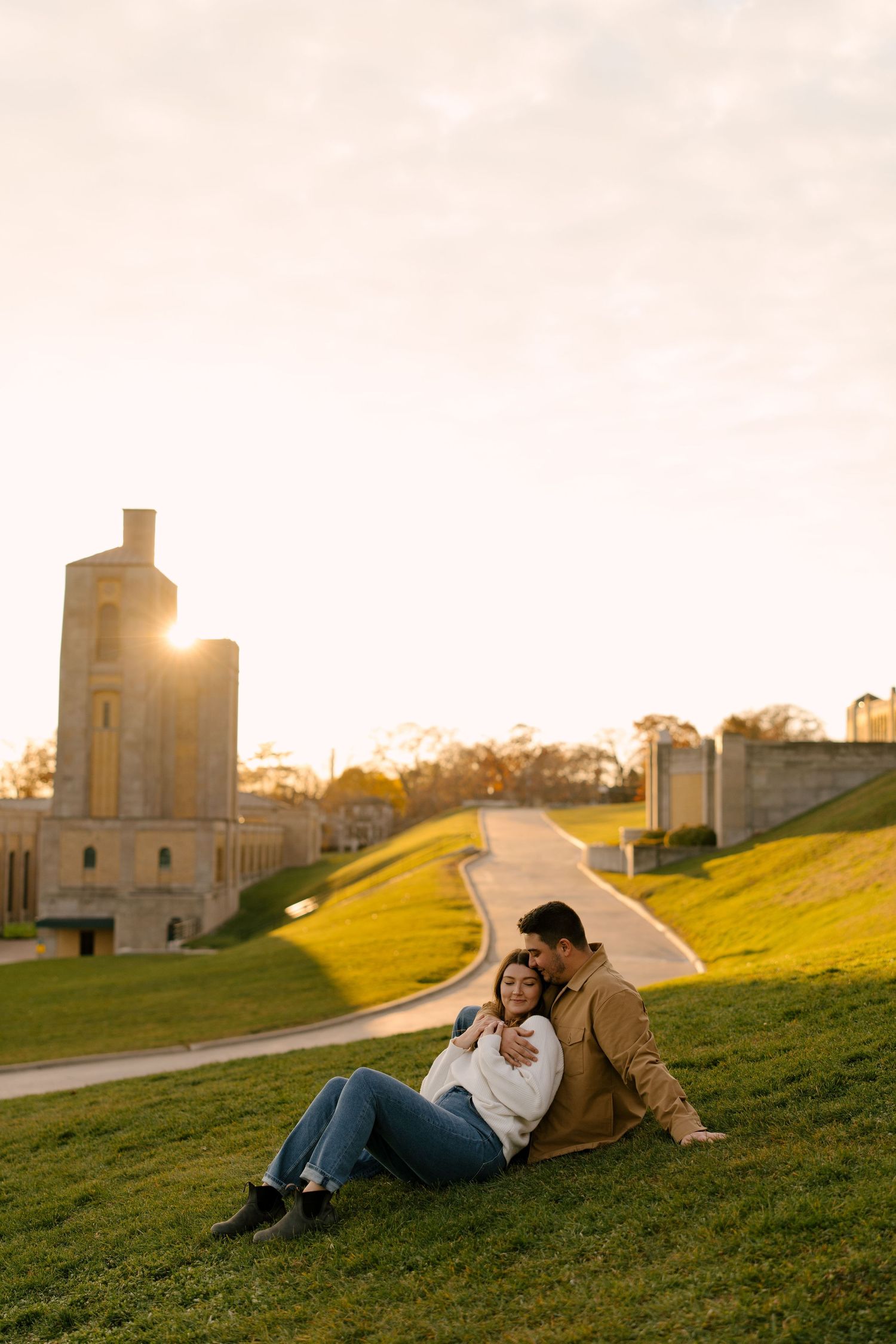 Engaged couple sitting together on a grassy hill at golden sunset with historic stone architecture behind.