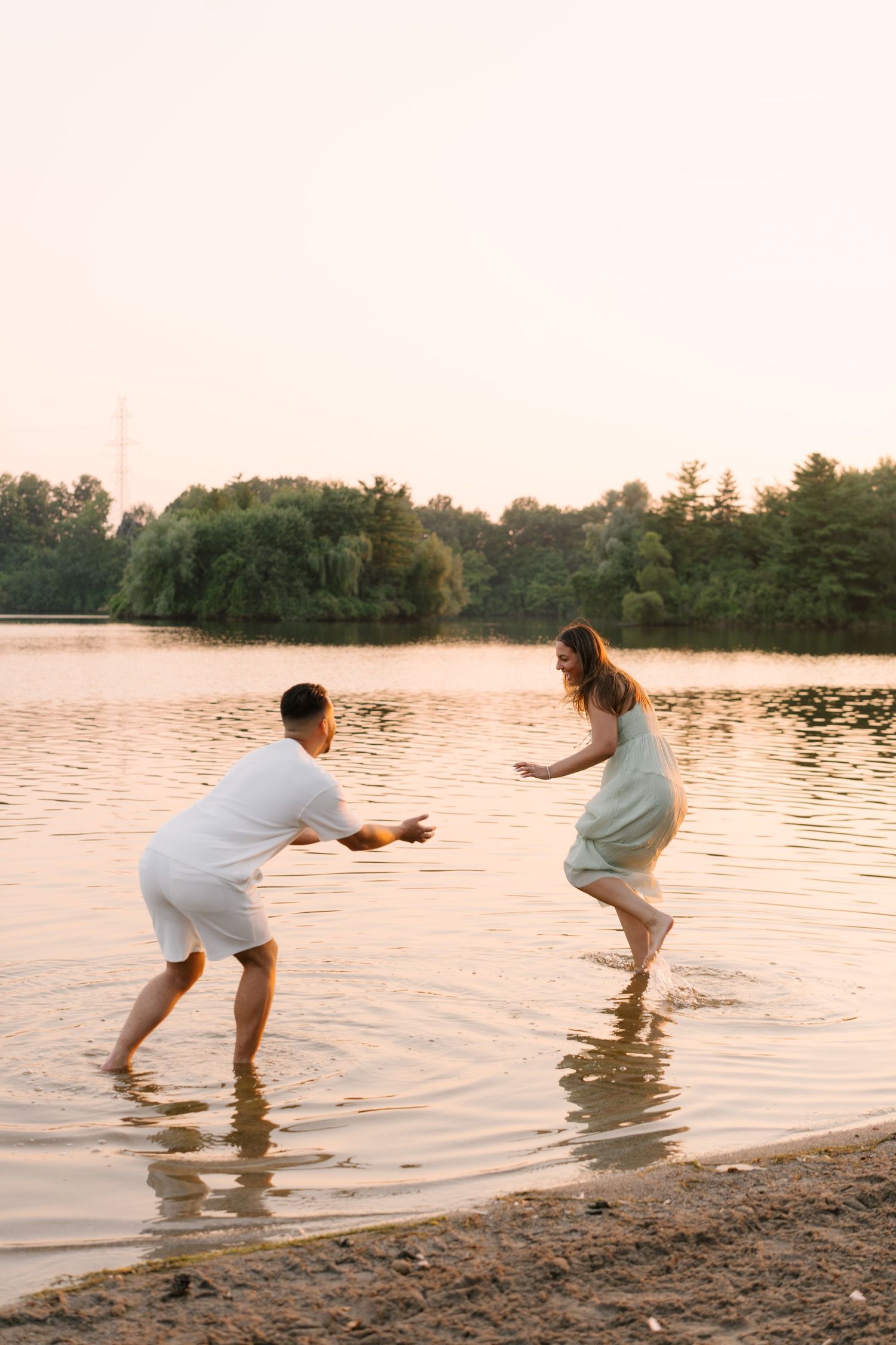 A playful couple splashes in shallow lake water at sunset, the man reaching out to catch the woman.