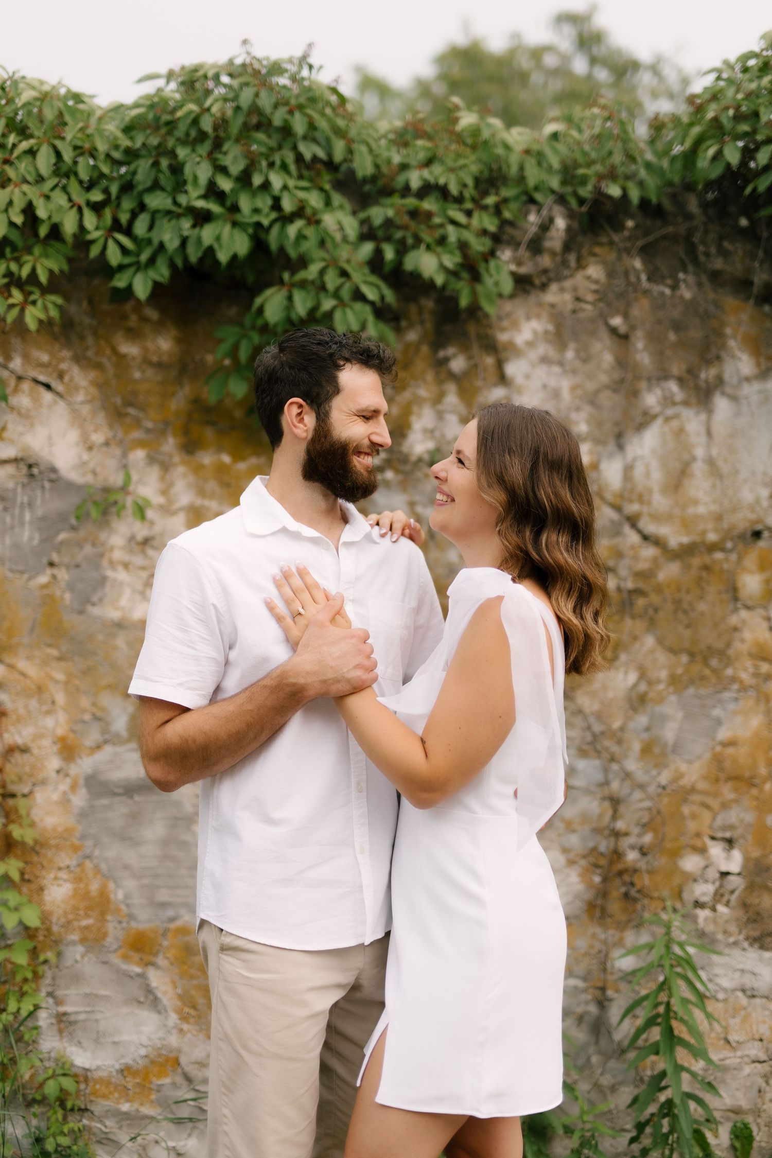 Smiling couple in white outfits embracing in front of a rustic stone wall covered in ivy.