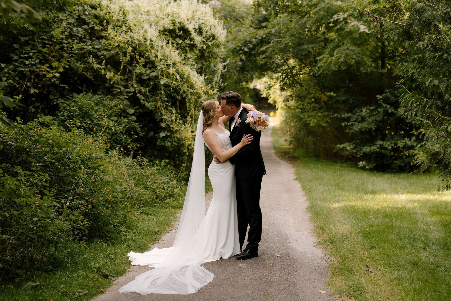 Bride and groom share a tender kiss on a tree-lined garden path on their wedding day.