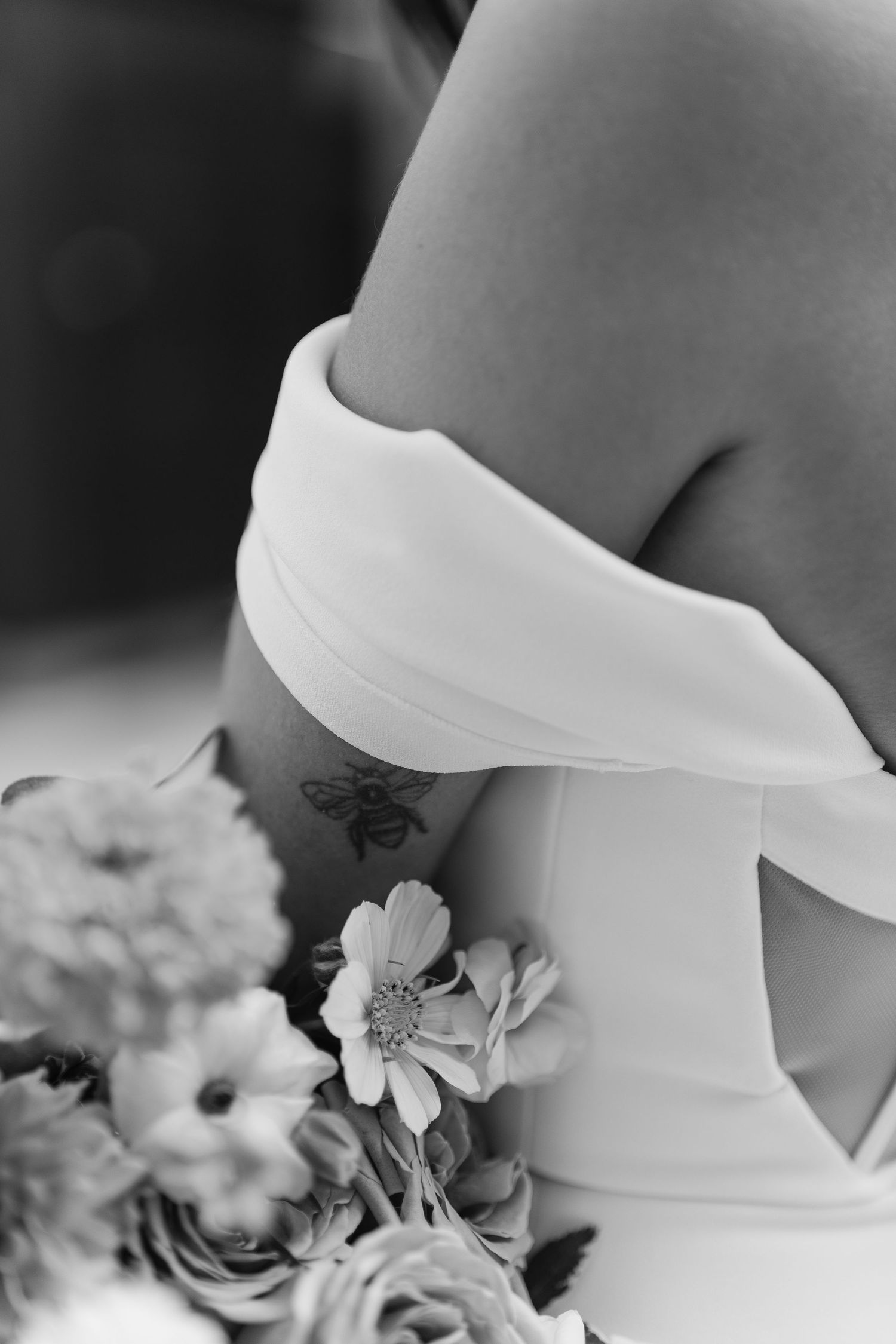 Close-up black and white photo of a bride in an off-shoulder gown holding a floral bouquet with a bee tattoo visible.