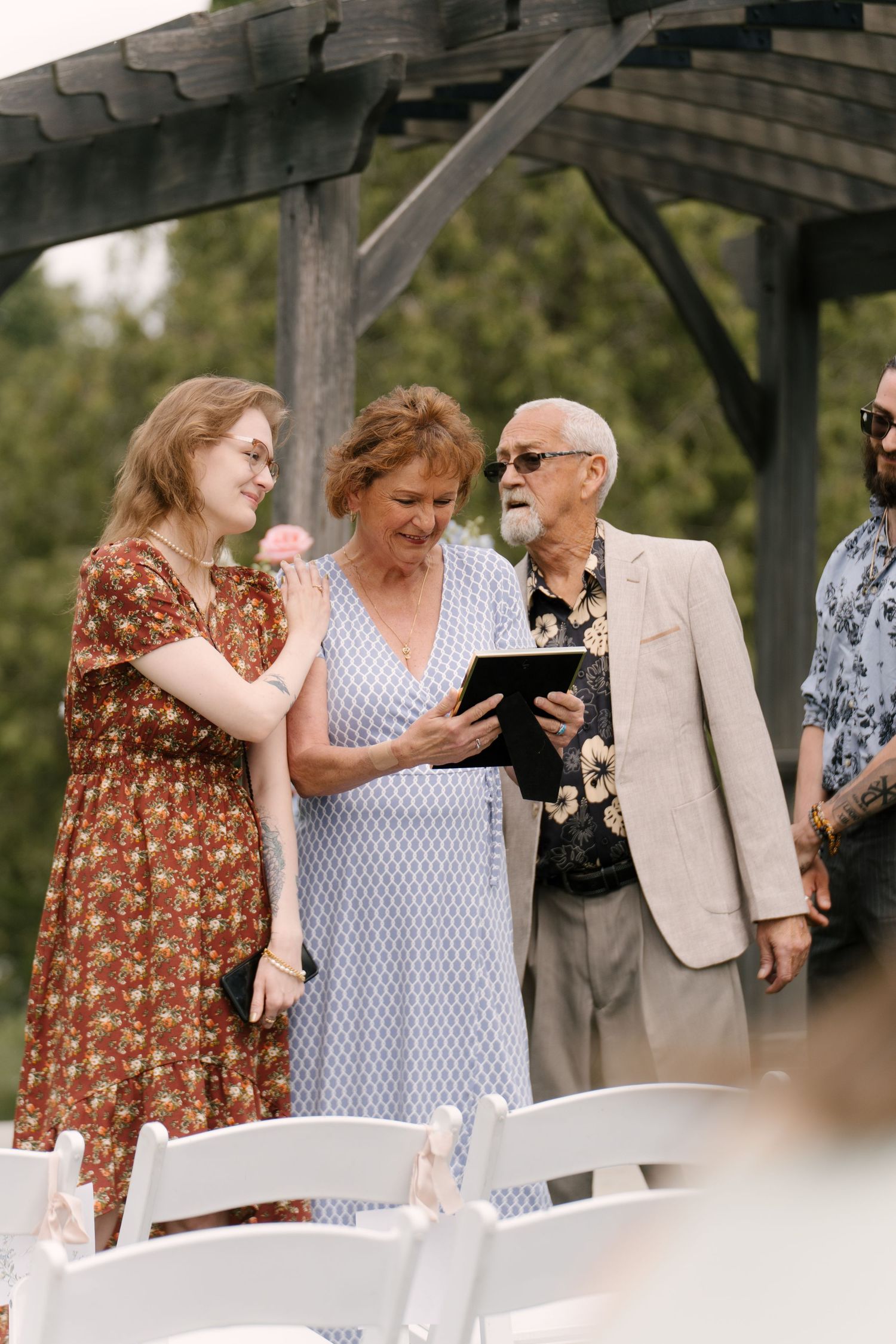 Wedding guests reviewing program under rustic pergola at outdoor garden ceremony.