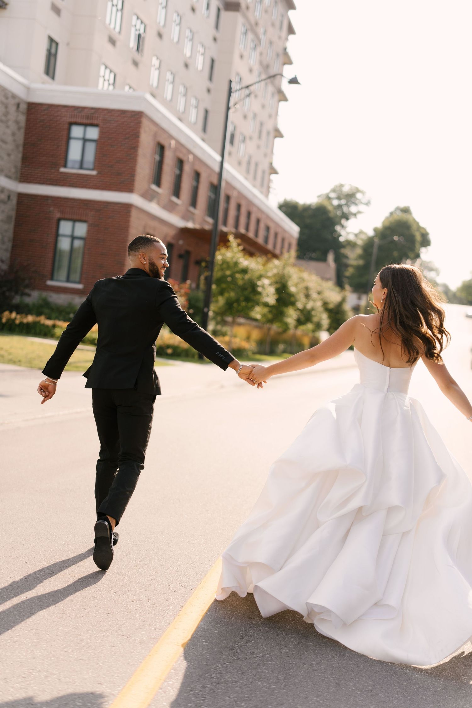Bride and groom hold hands running down street, bride in ruffled white gown, groom in black suit outside Tapestry Hall venue.