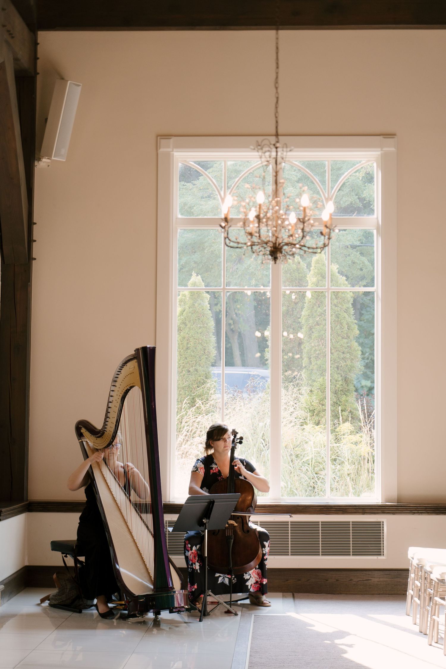 Two musicians playing harp and cello in an elegant Ancaster Mill chapel room with chandelier and large window.