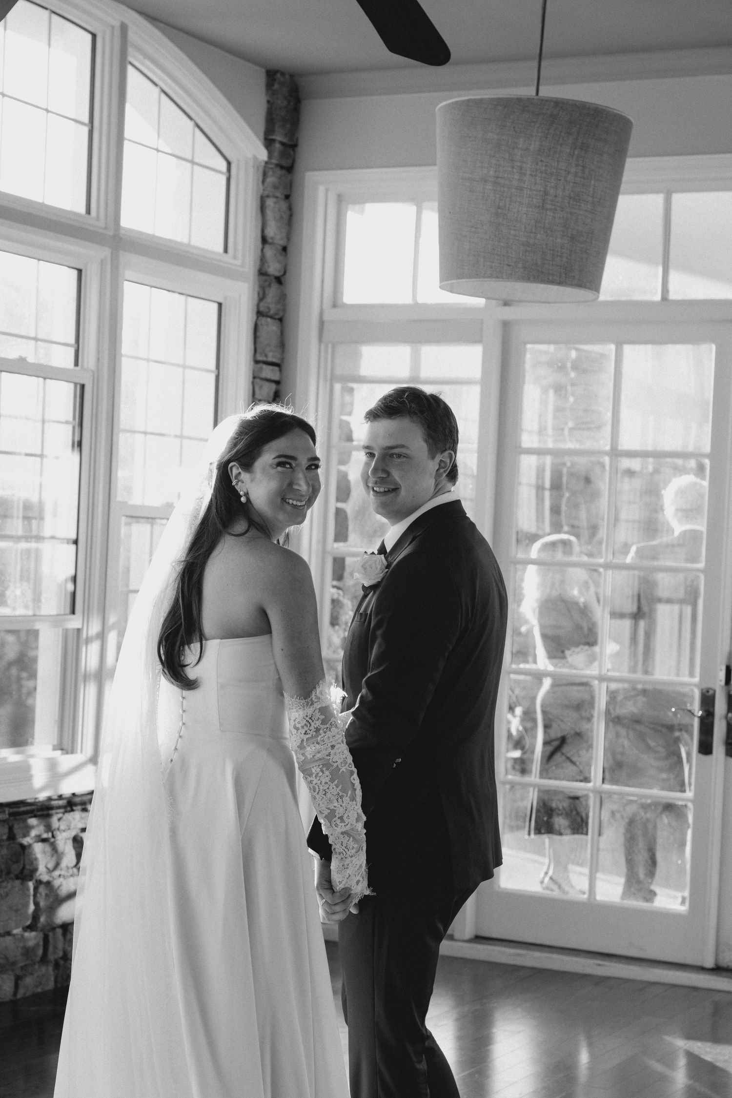 Black and white photo of smiling bride and groom standing indoors near large windows on wedding day.