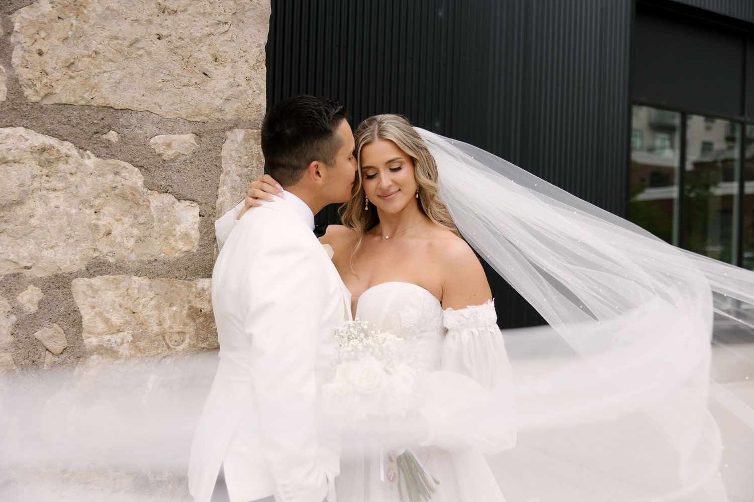 Groom in white suit kisses bride's cheek as her veil flows in the wind outside Tapestry Hall venue.