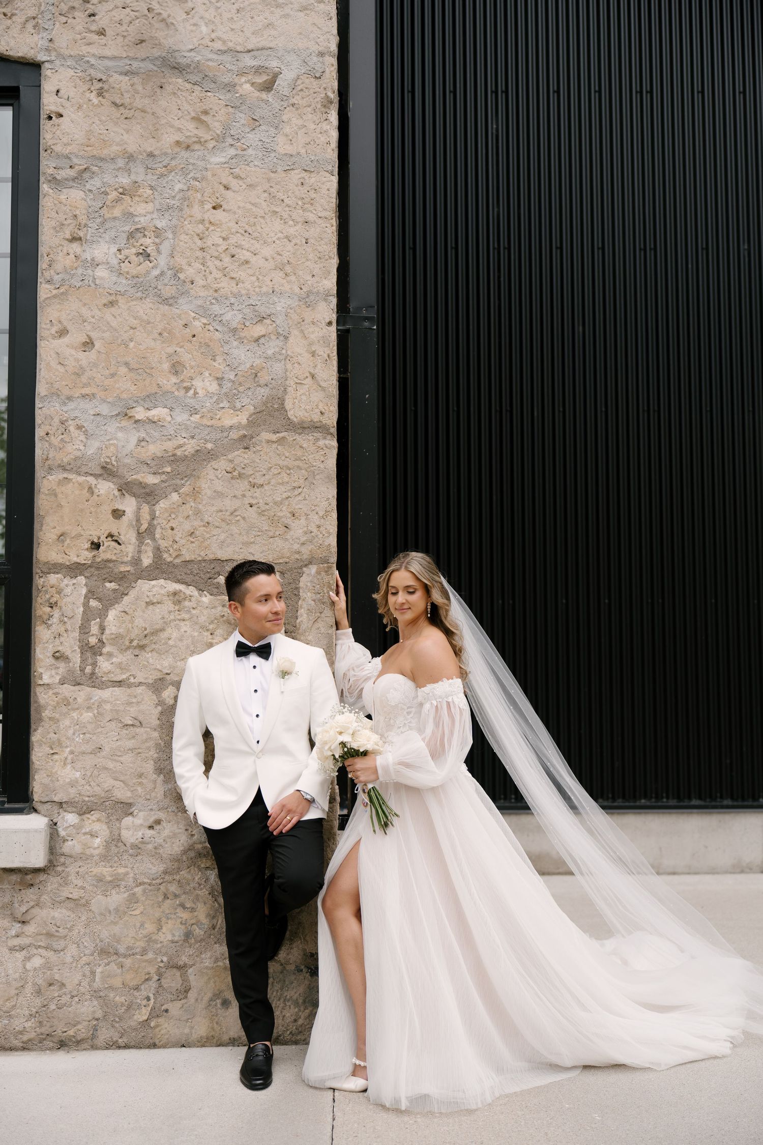 Bride and groom pose against a stone wall and black barn door in elegant wedding attire at Tapestry Hall wedding venue.
