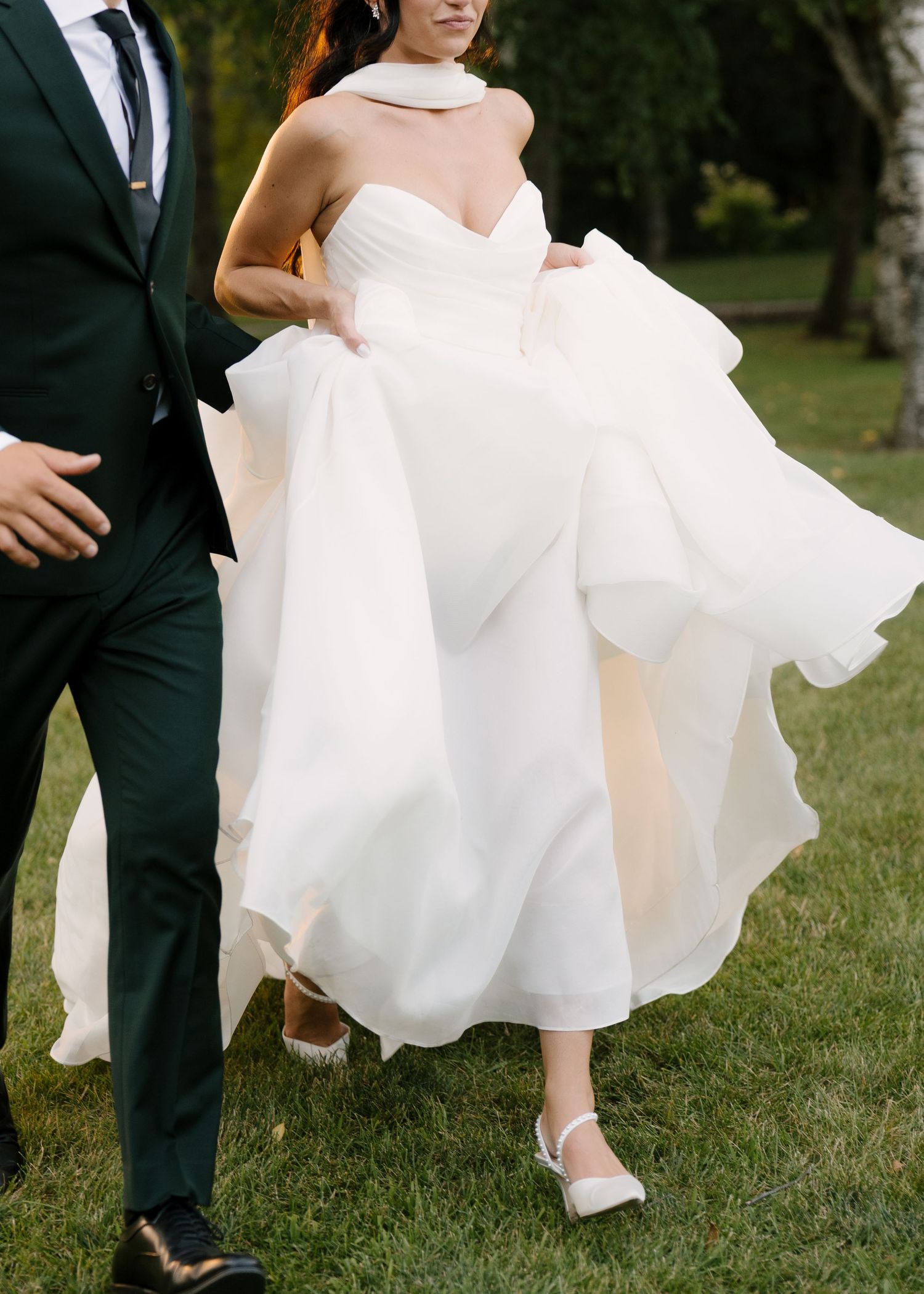 Bride lifting flowing white ball gown skirt while walking outdoors, groom in dark green suit beside her.