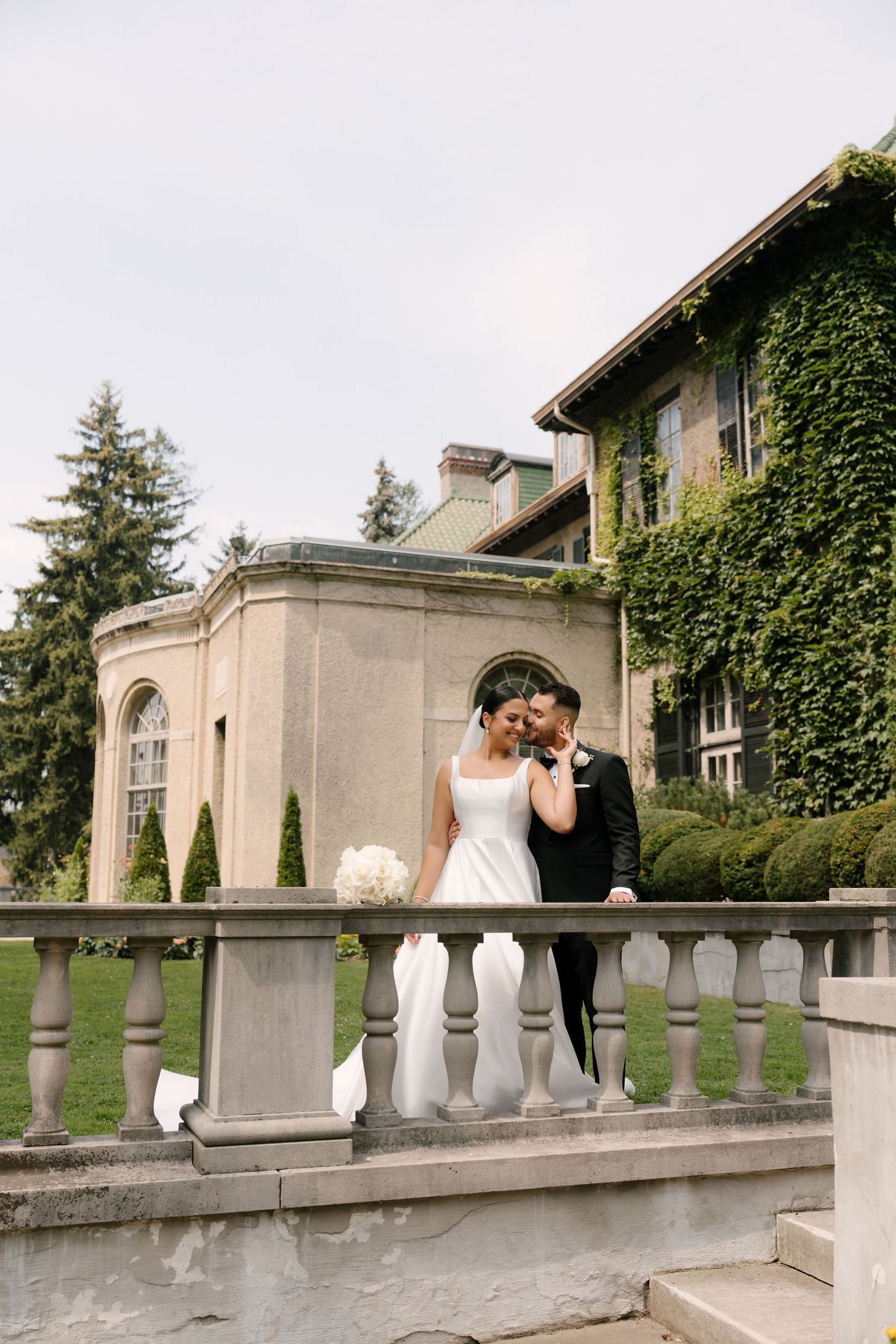 Bride and groom sharing a kiss on a stone balustrade terrace outside a grand ivy-covered estate wedding venue.