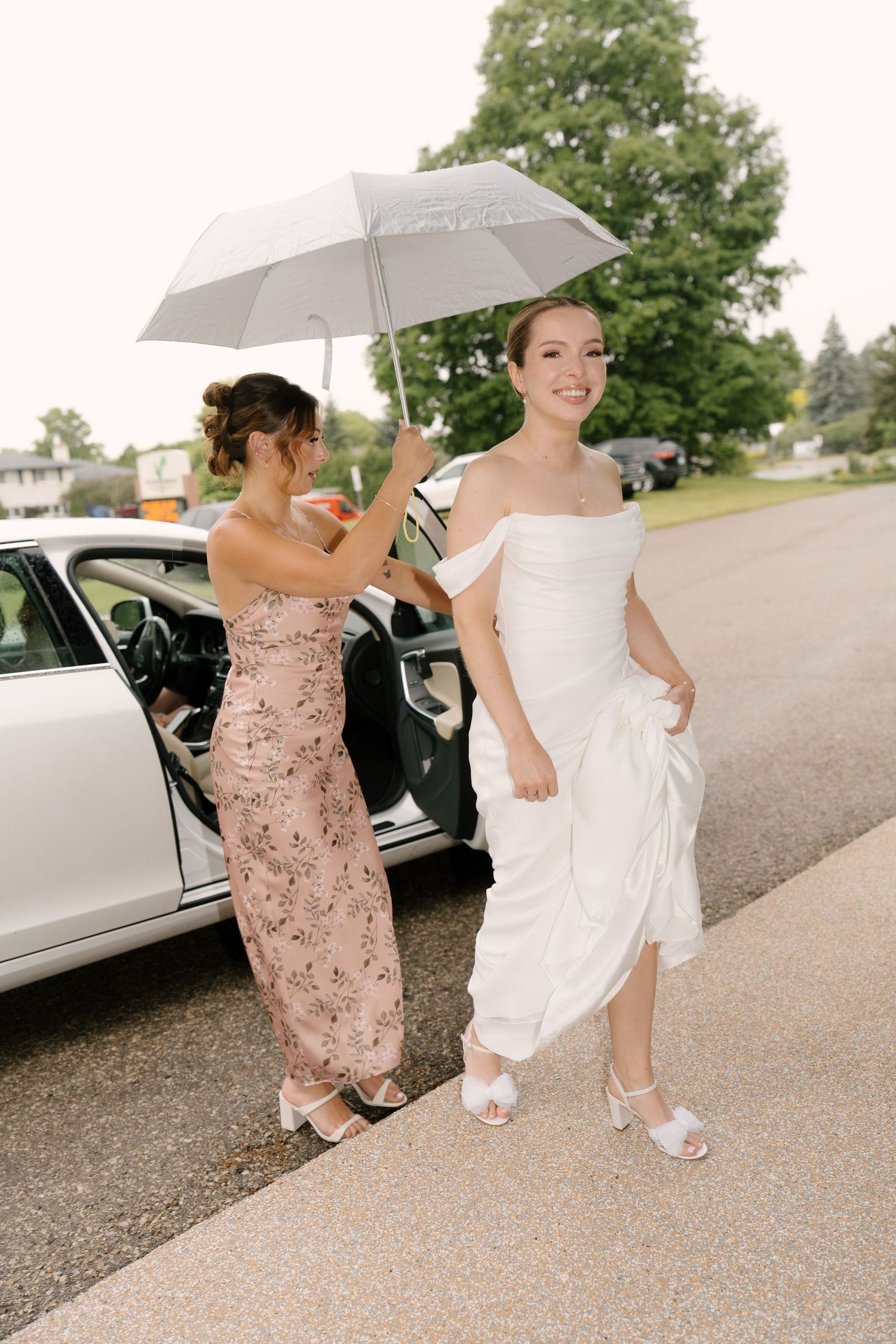 Bridesmaid holding white umbrella over smiling bride arriving at wedding venue from car.