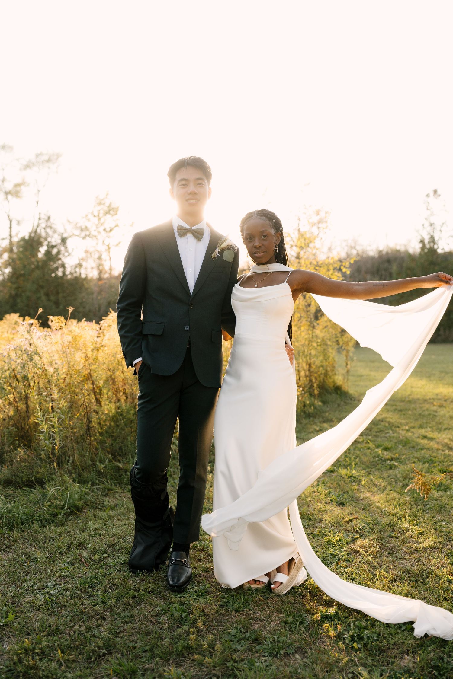 A bride and groom pose together at sunset in a field, her white veil flowing gracefully in the breeze at Wren House venue.