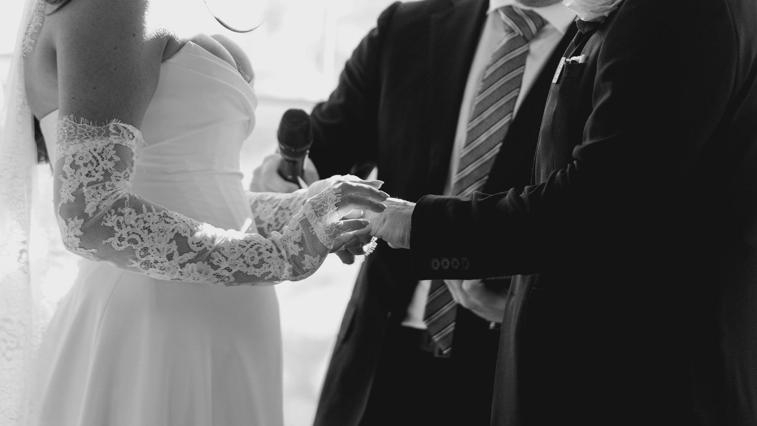 Black and white photo of groom placing ring on bride's lace-sleeved hand at altar.