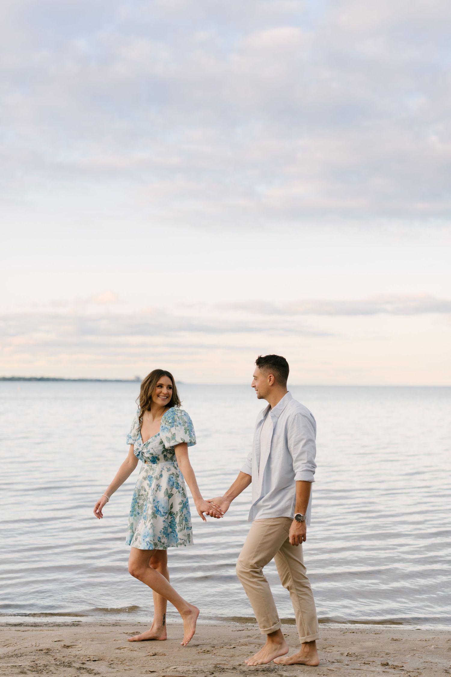 Couple holding hands and walking barefoot along a sandy beach at sunset with calm water behind them.