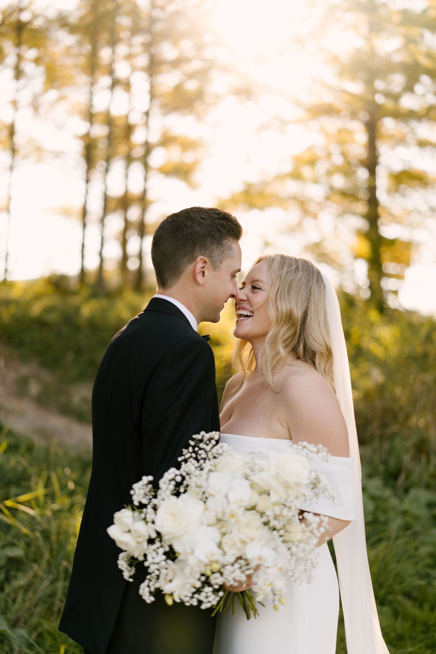 Bride and groom share a romantic nose-to-nose moment outdoors with golden sunlight filtering through trees.