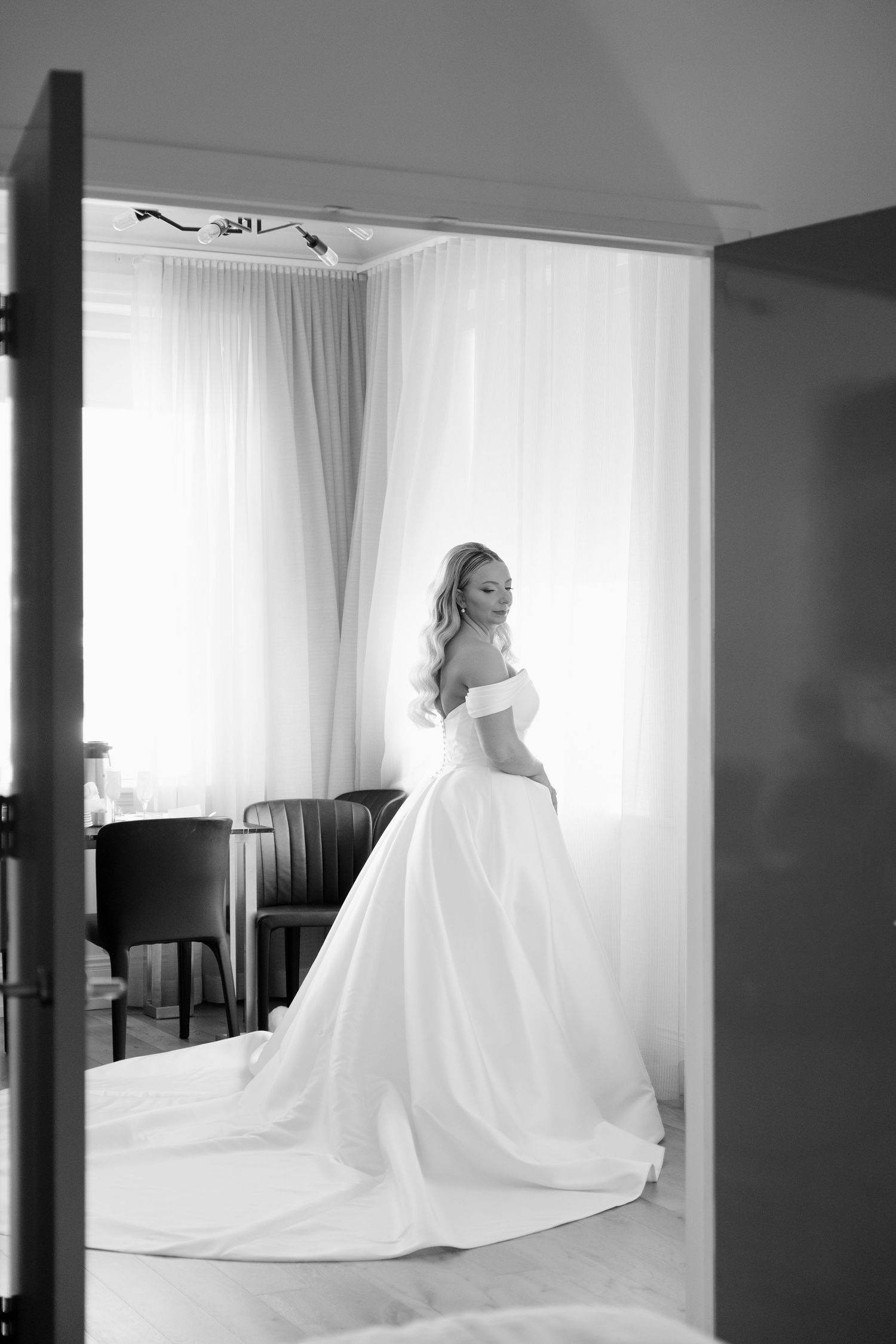 Black and white portrait of elegant bride in off-shoulder wedding gown standing by bright window in The Walper Hotel.