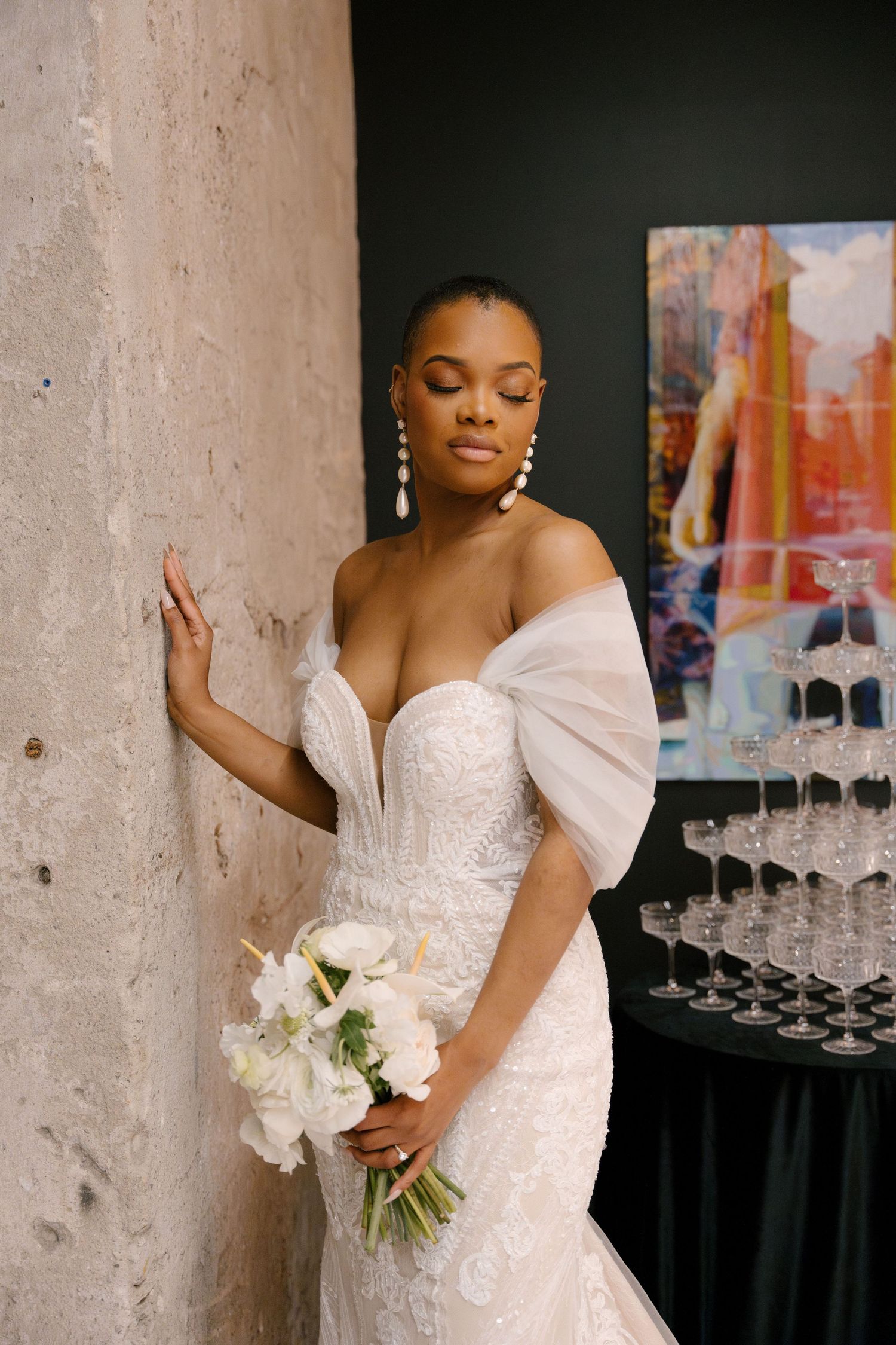 Elegant bride in strapless lace gown holding white bouquet, leaning against stone wall near champagne tower.