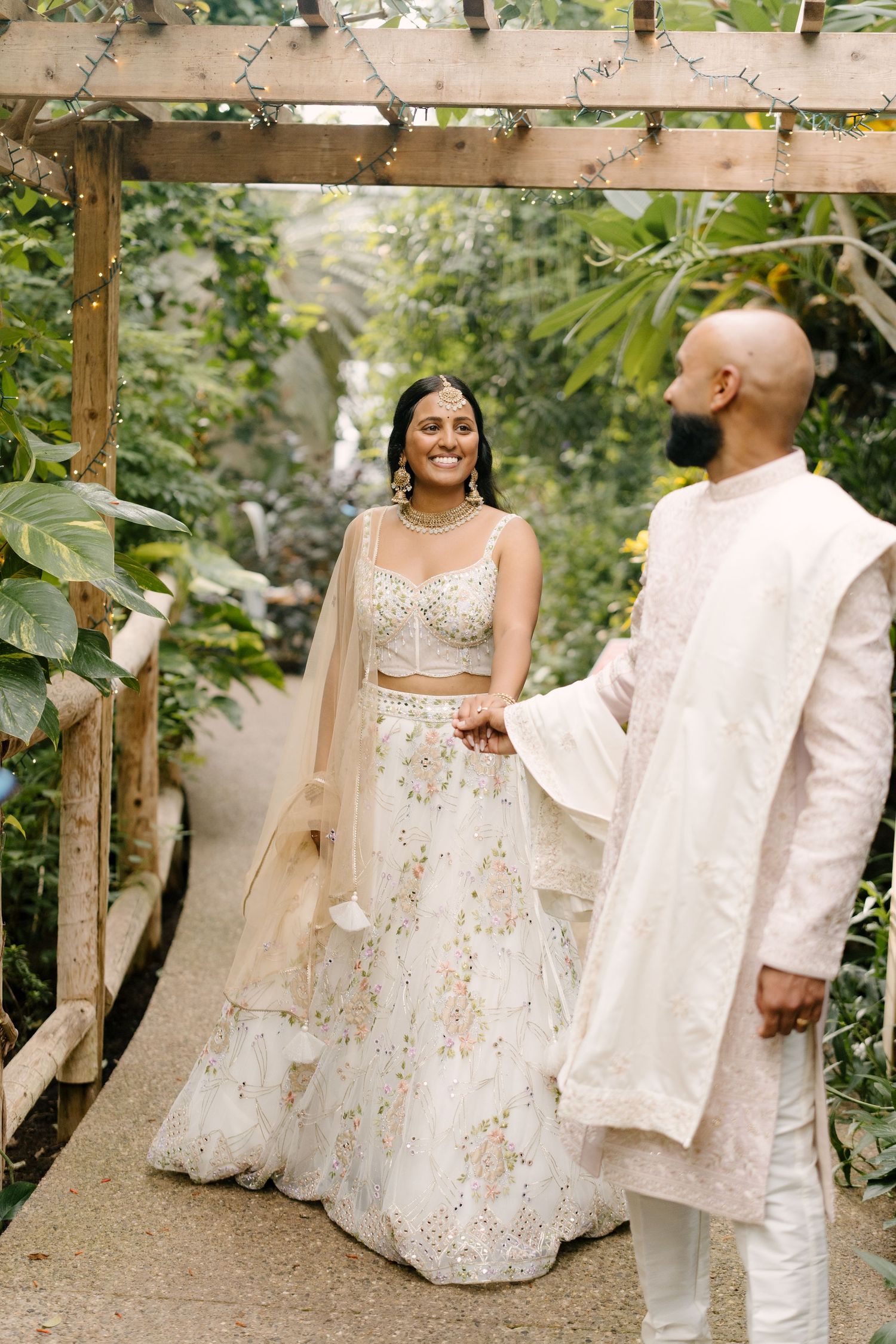 Smiling Indian bride in embroidered lehenga holds hands with groom in white.