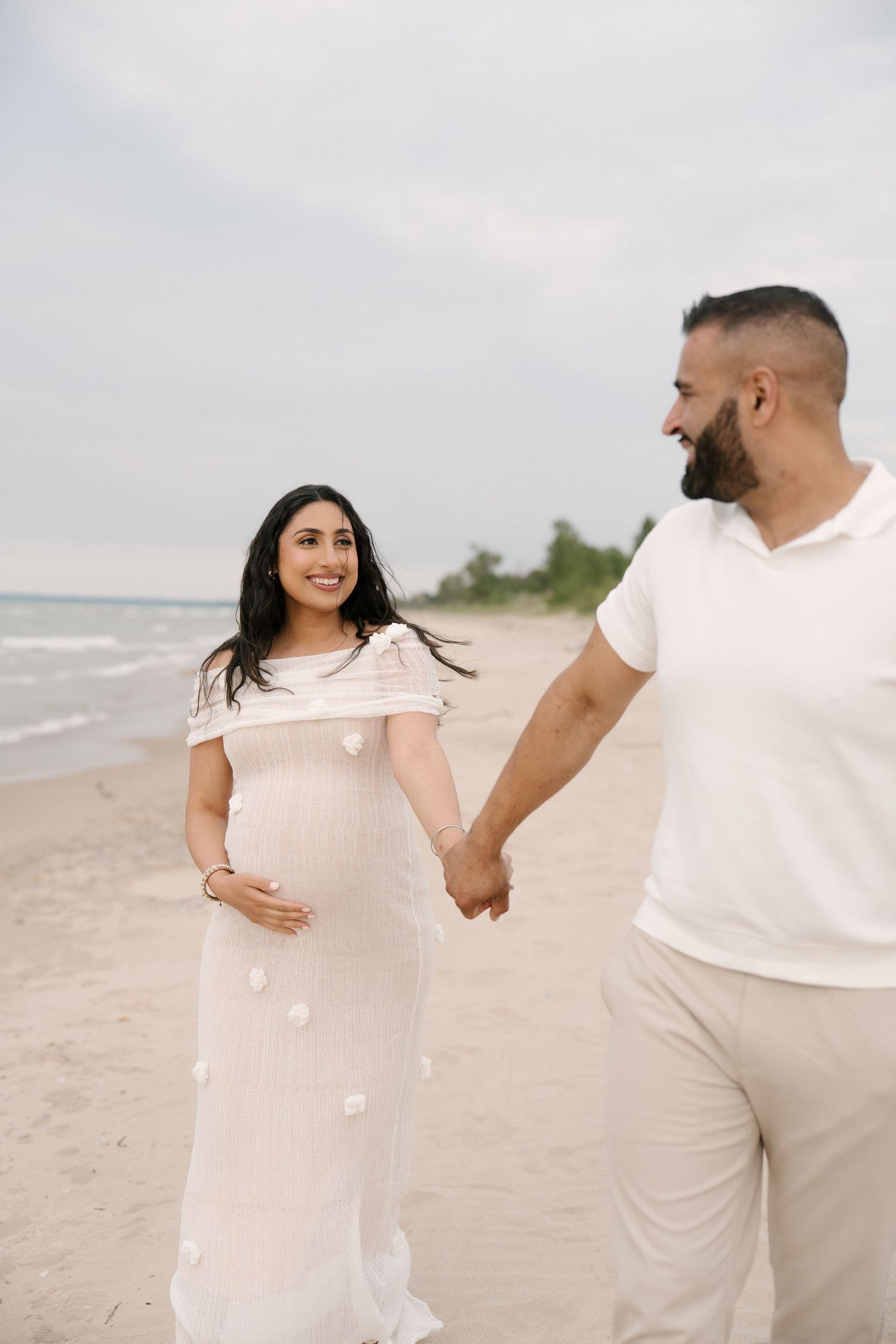 Smiling pregnant woman in white floral dress holds partners hand while walking on sandy beach.