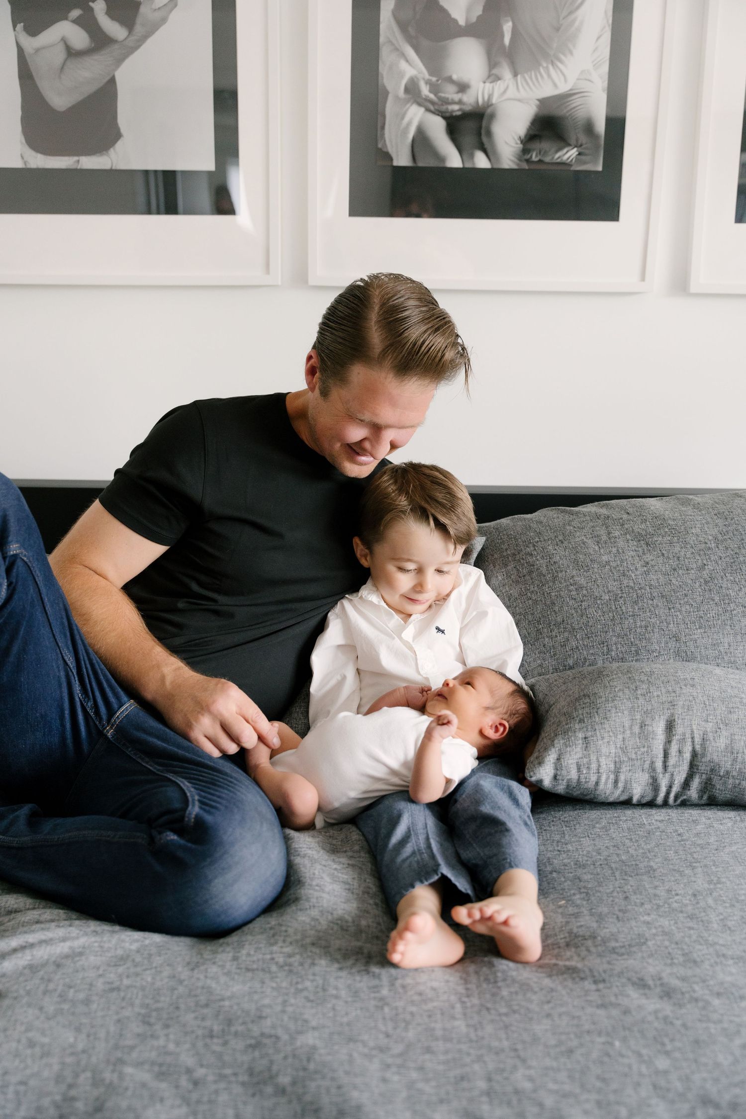 Father in black shirt sitting on gray couch with toddler son holding newborn baby gently.
