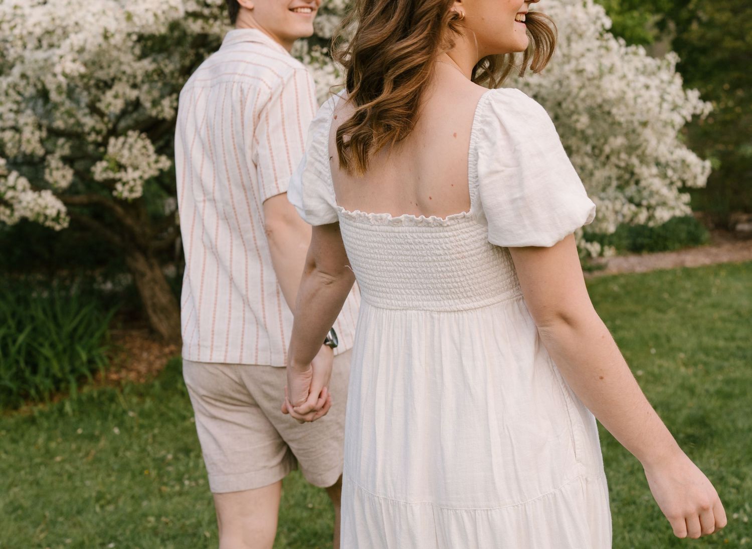 Couple holding hands and walking together in a lush garden with white flowering trees in the background.