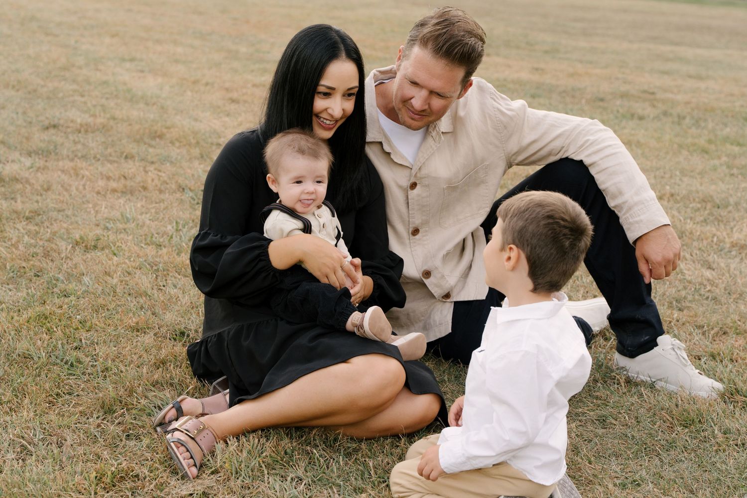 Happy family of four sits together on a grassy field, smiling and enjoying quality time outdoors.
