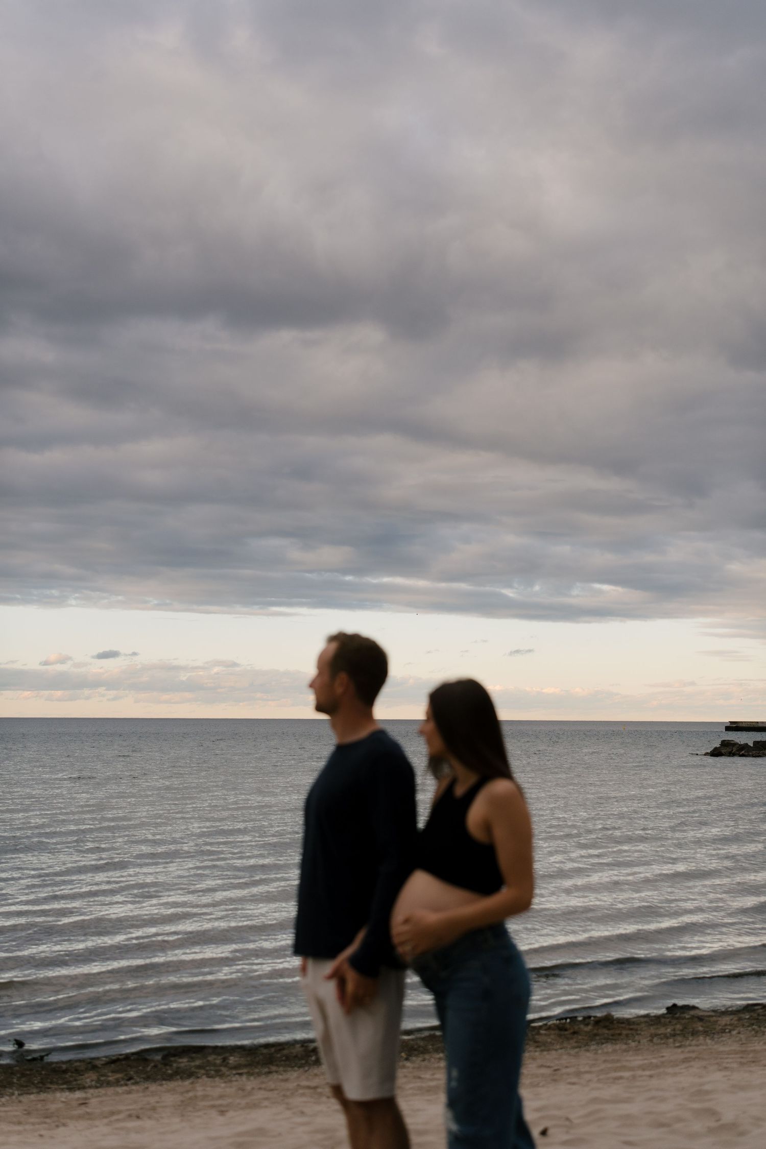 Expecting couple stands together on beach at sunset, woman cradling her pregnant belly near calm ocean waters.