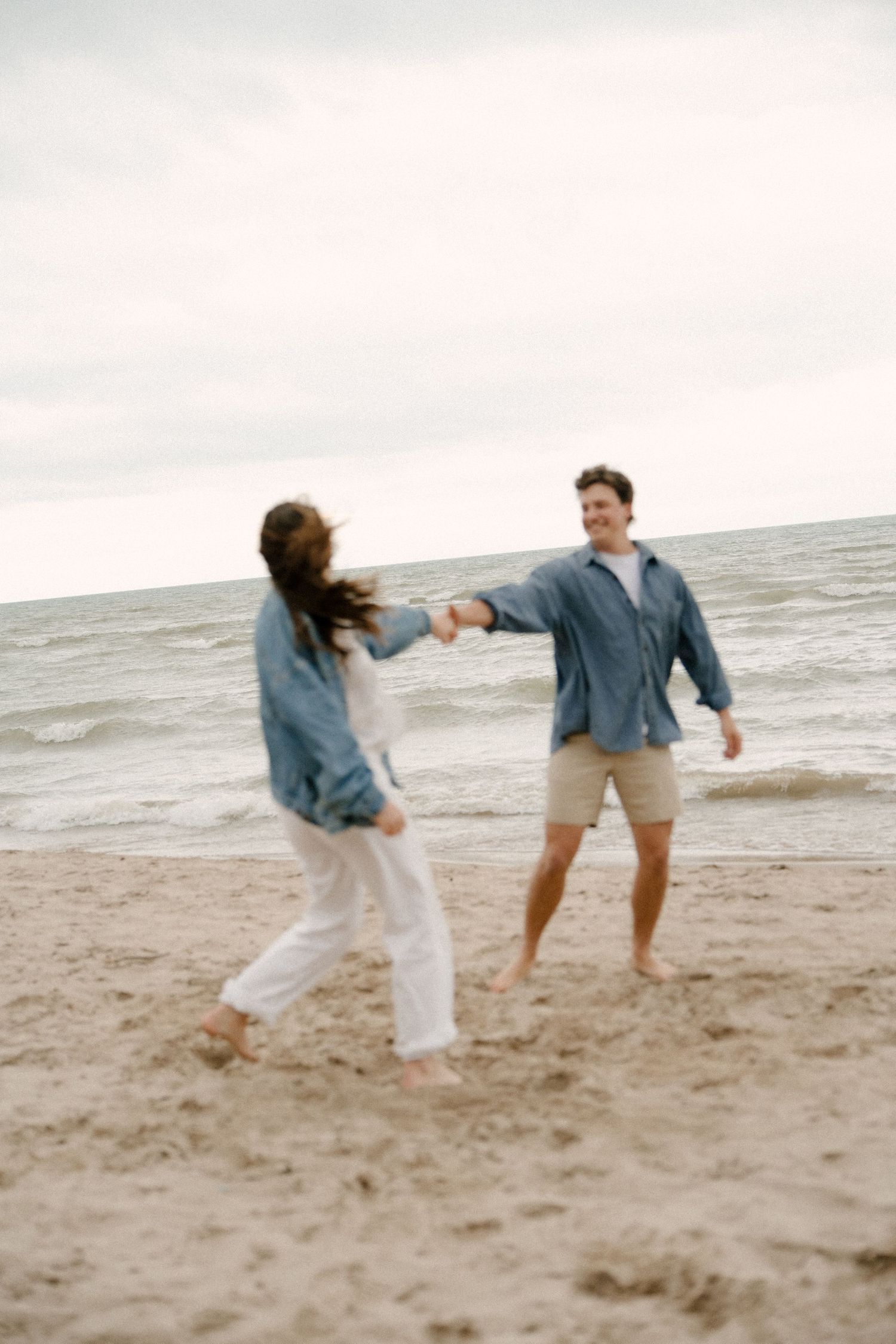Couple holding hands and dancing playfully together on a sandy beach with ocean waves behind them.