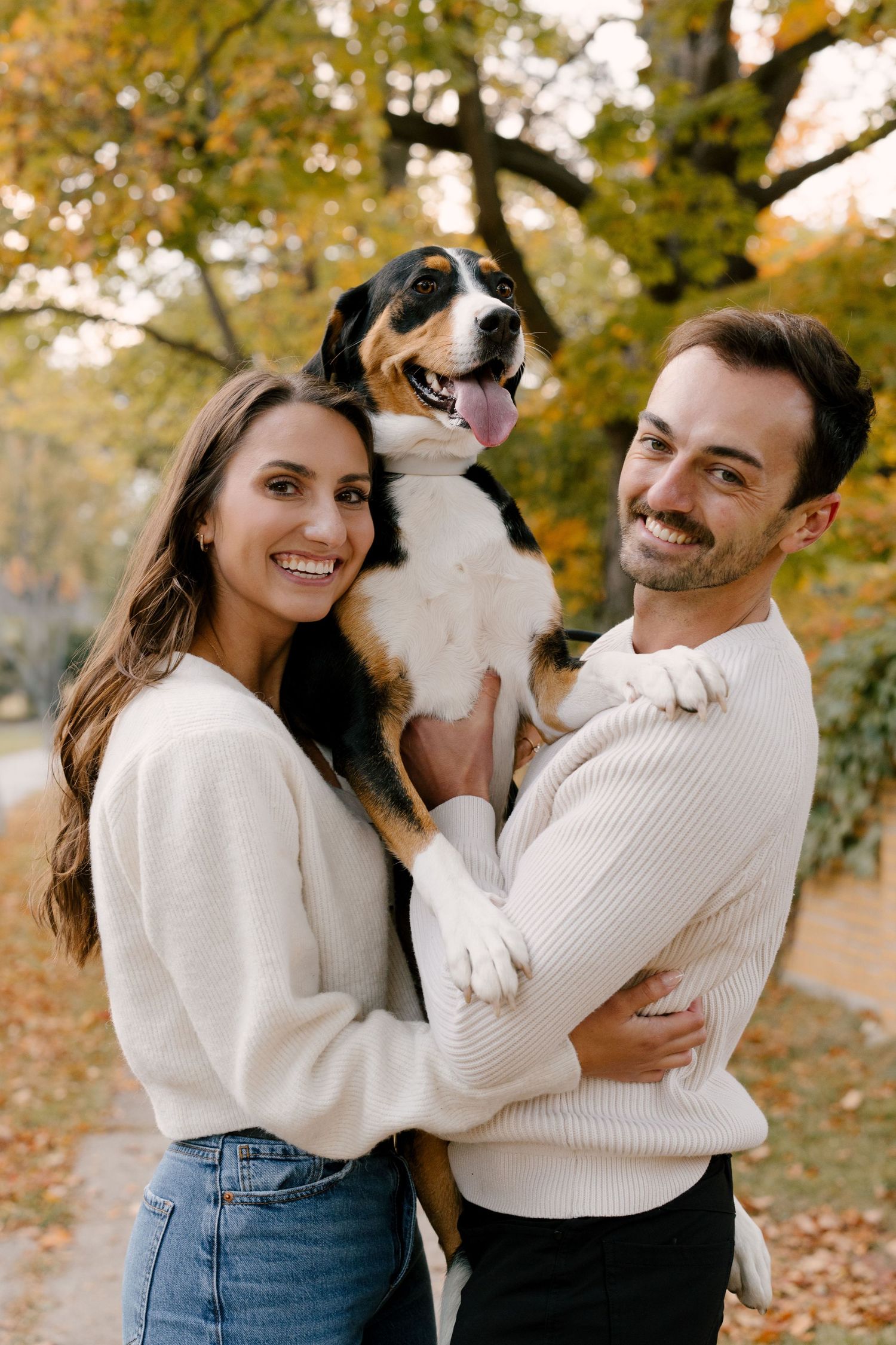 Smiling couple holding their Bernese mix dog in a colorful autumn park setting.