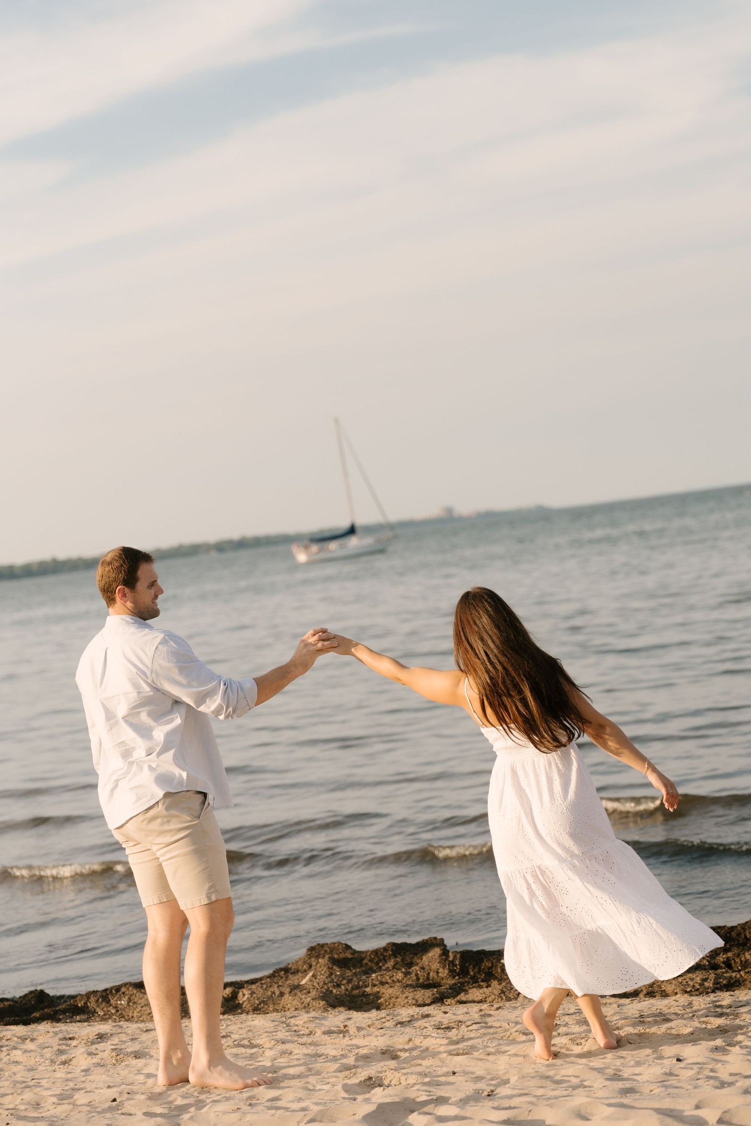 Couple dancing playfully on sandy beach near ocean, woman in white dress, sailboat visible in background.
