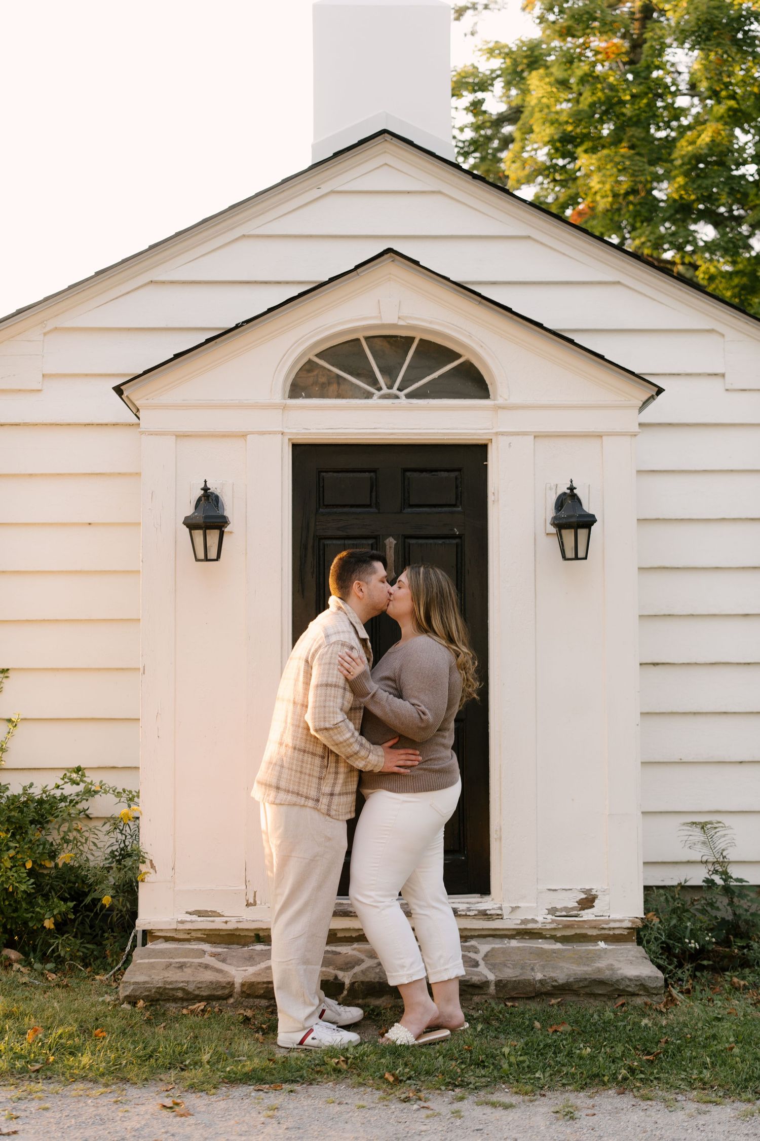 Couple sharing a kiss in front of charming white colonial building entrance.