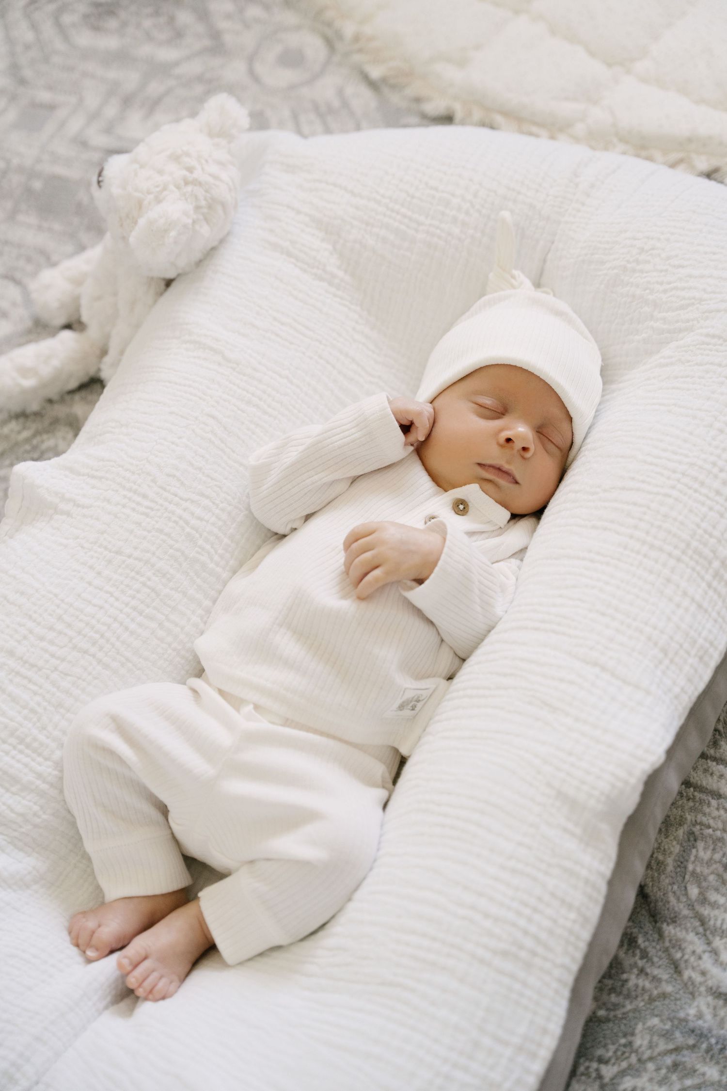 Sleeping newborn in white knit outfit and hat resting on cream pillow.