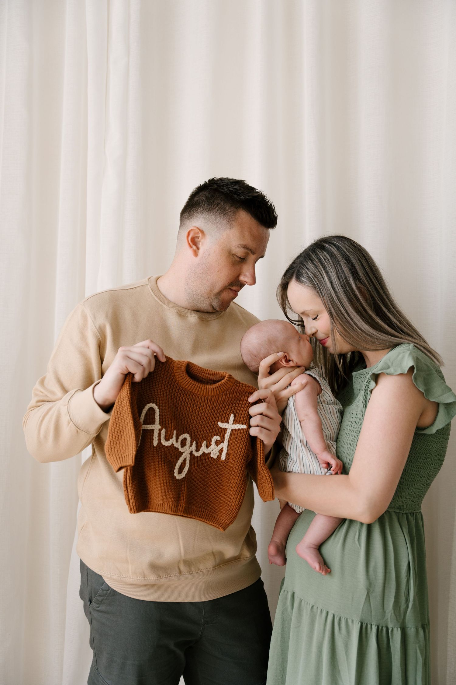 Parents lovingly gaze at their newborn while father holds a brown knit onesie reading August.
