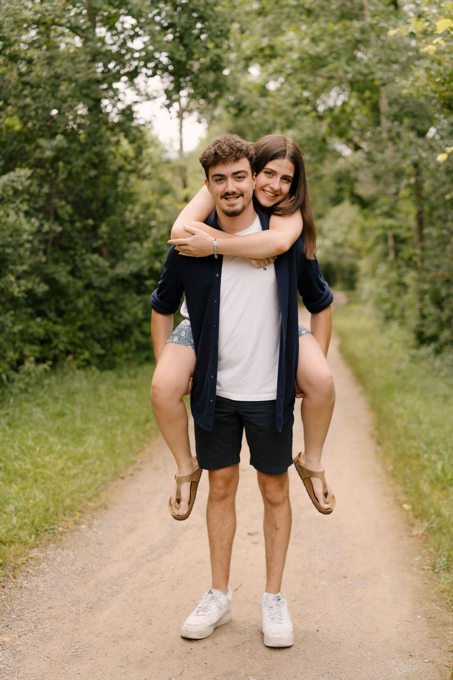Happy young couple on a forest path, man giving woman a piggyback ride, smiling outdoors in summer.