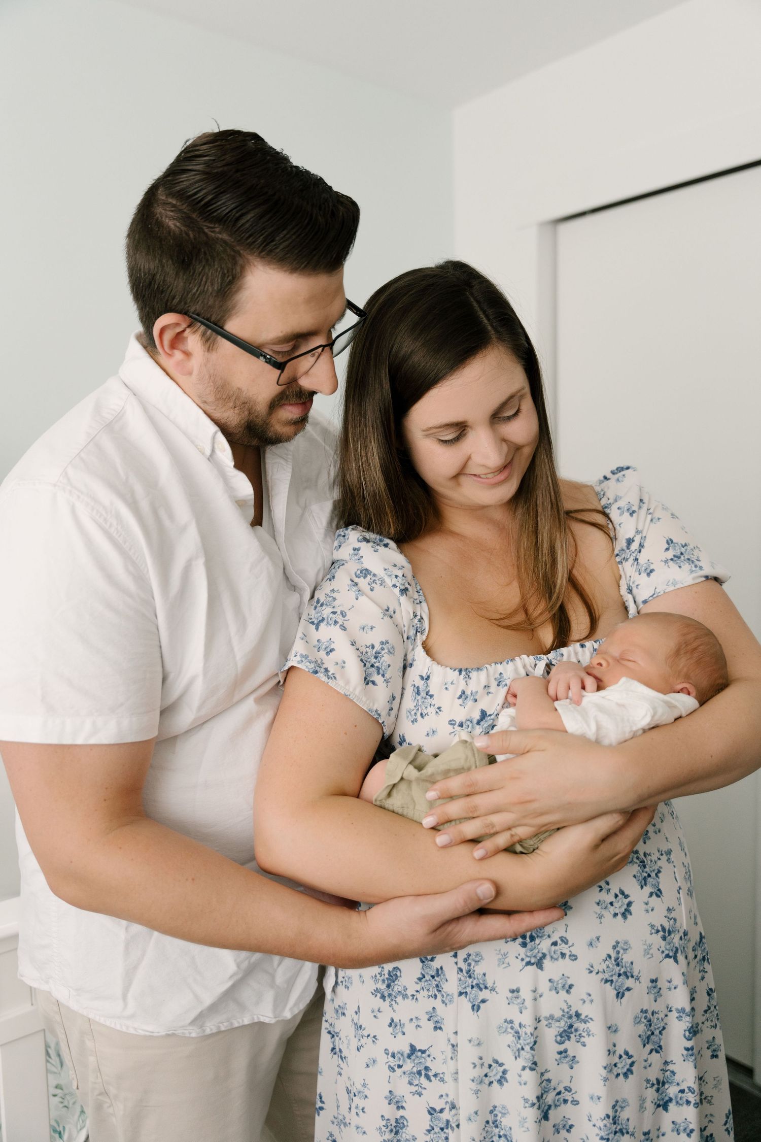 Smiling new parents with glasses standing together in bright room holding their sleeping newborn baby.