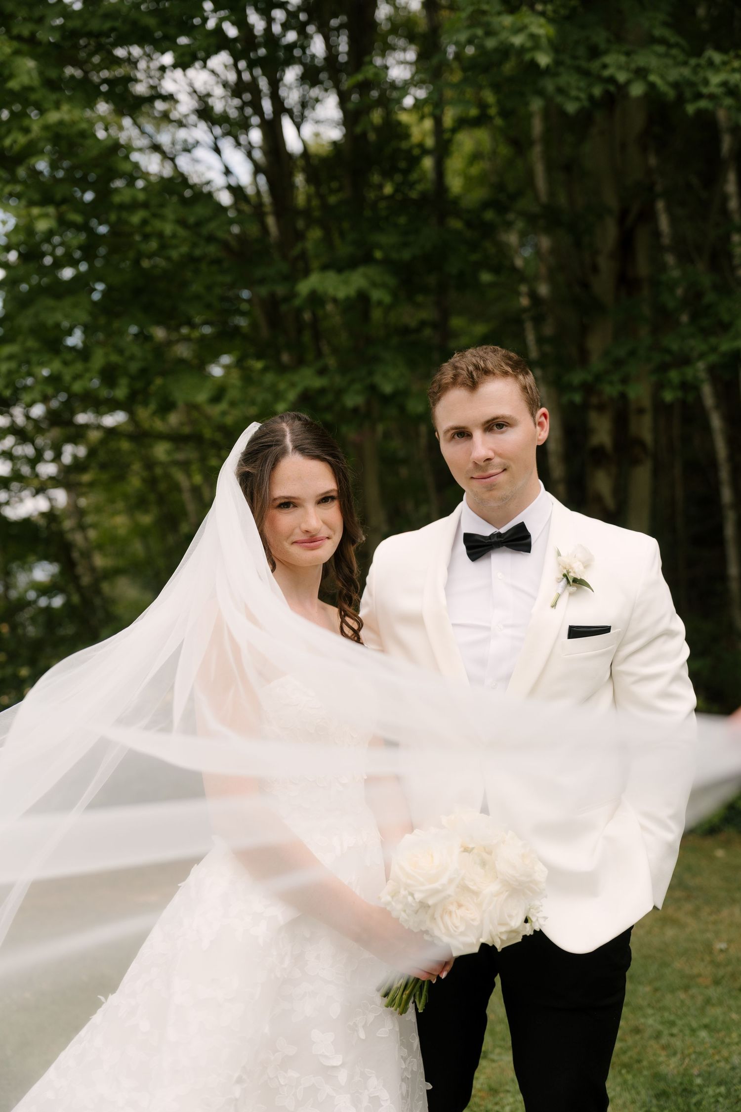 Bride and groom pose outdoors with flowing veil, white bouquet, and elegant attire against lush green trees.